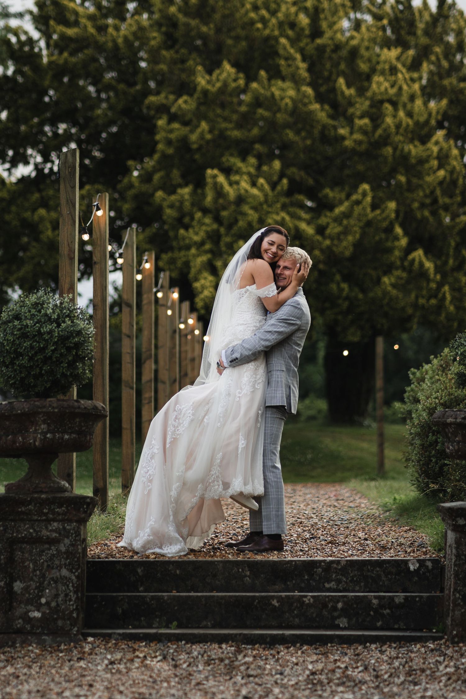 A romantic wedding couple shares an intimate moment on stone steps surrounded by string lights and greenery at dusk.