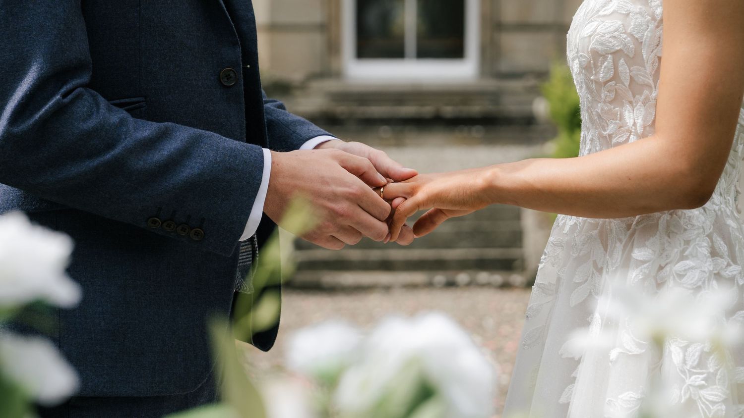 Close-up of hands exchanging wedding rings during outdoor ceremony with white lace dress and navy suit visible.