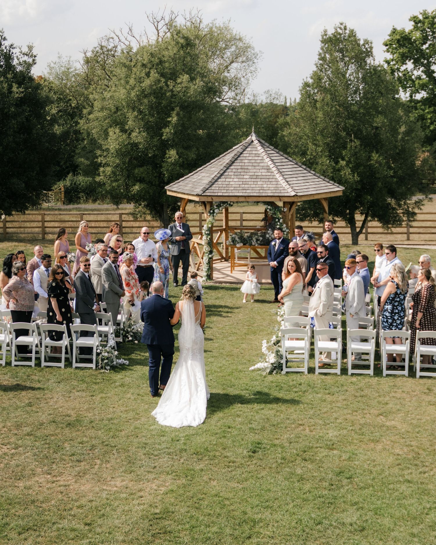 A wedding ceremony takes place outdoors with guests seated on white chairs and a gazebo in the background.
