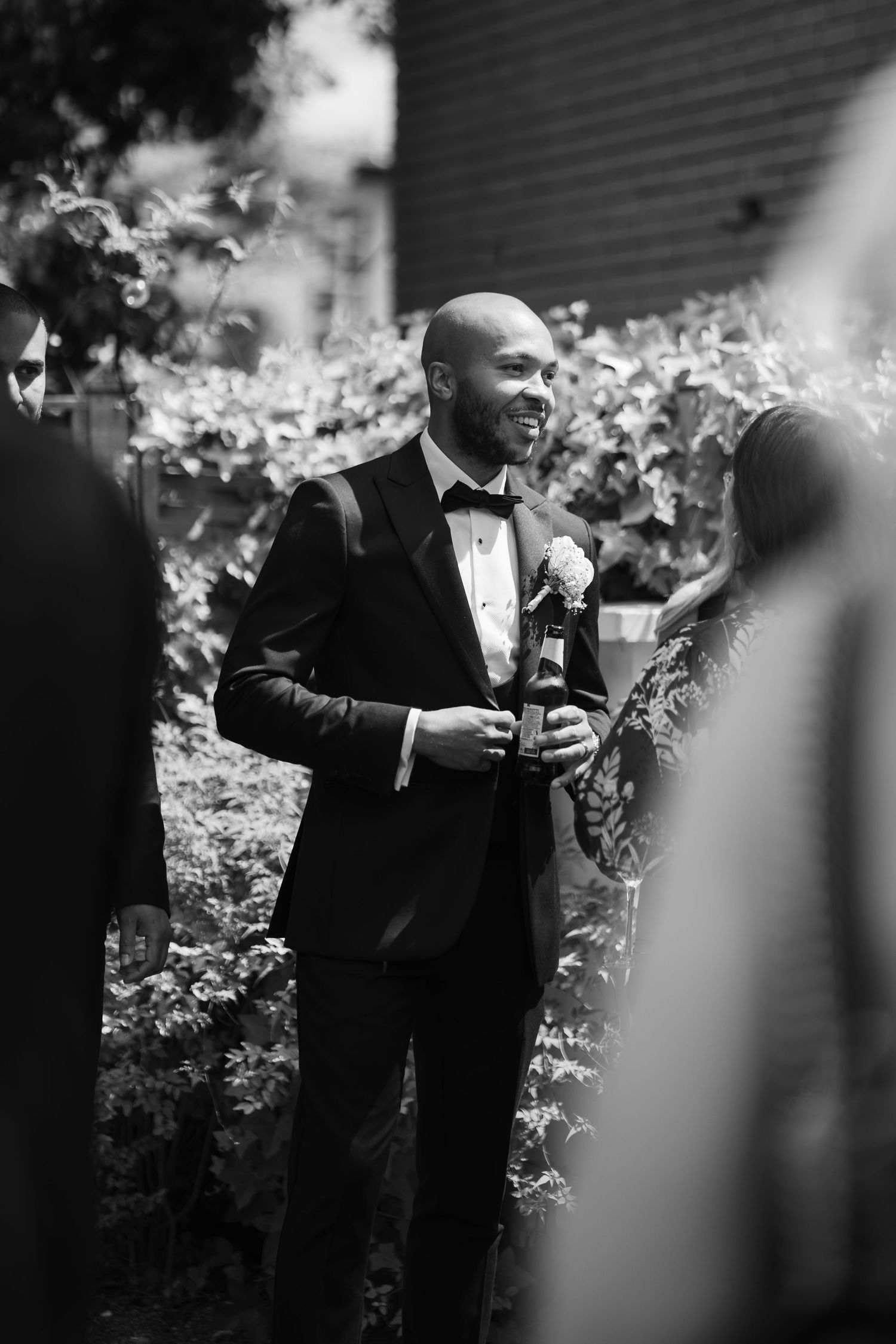 A person in a tuxedo stands in front of white flowers while looking at their phone in black and white photography.