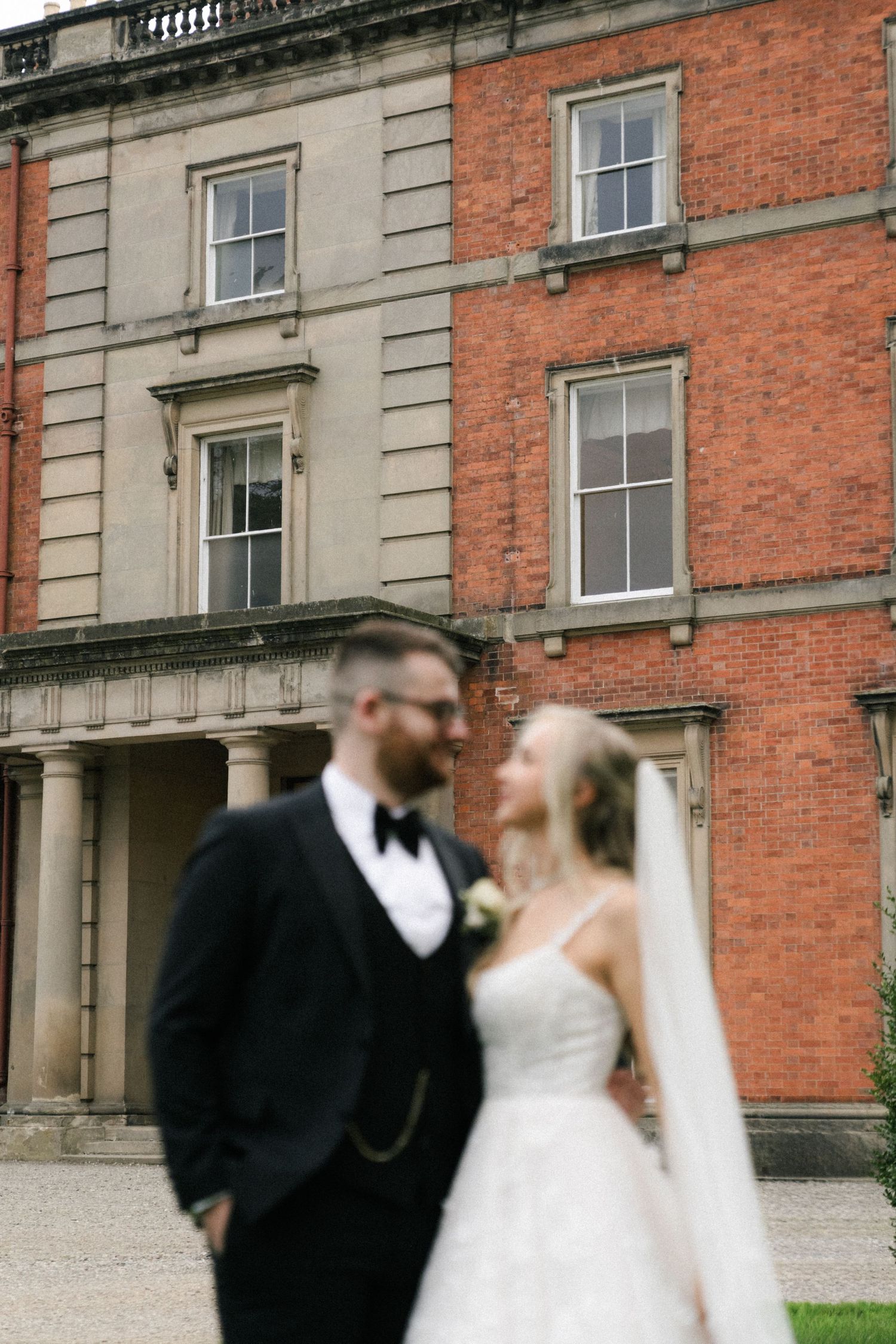 A wedding couple shares a romantic moment outside a grand red brick mansion with classical architecture.
