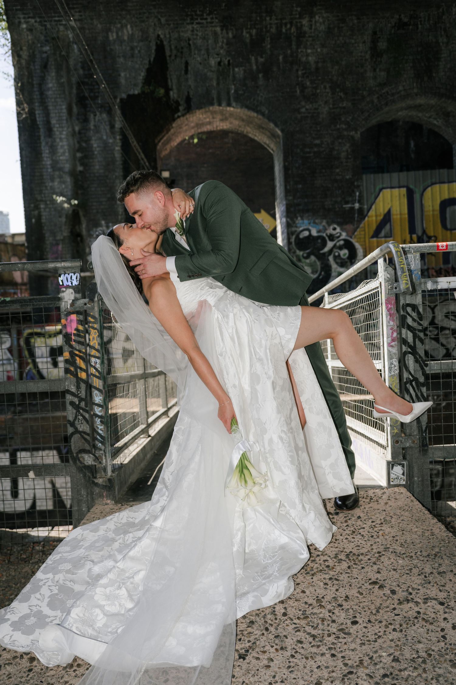 A bride in a white wedding dress lies dramatically on a pile of gravel in an urban setting with graffiti walls.