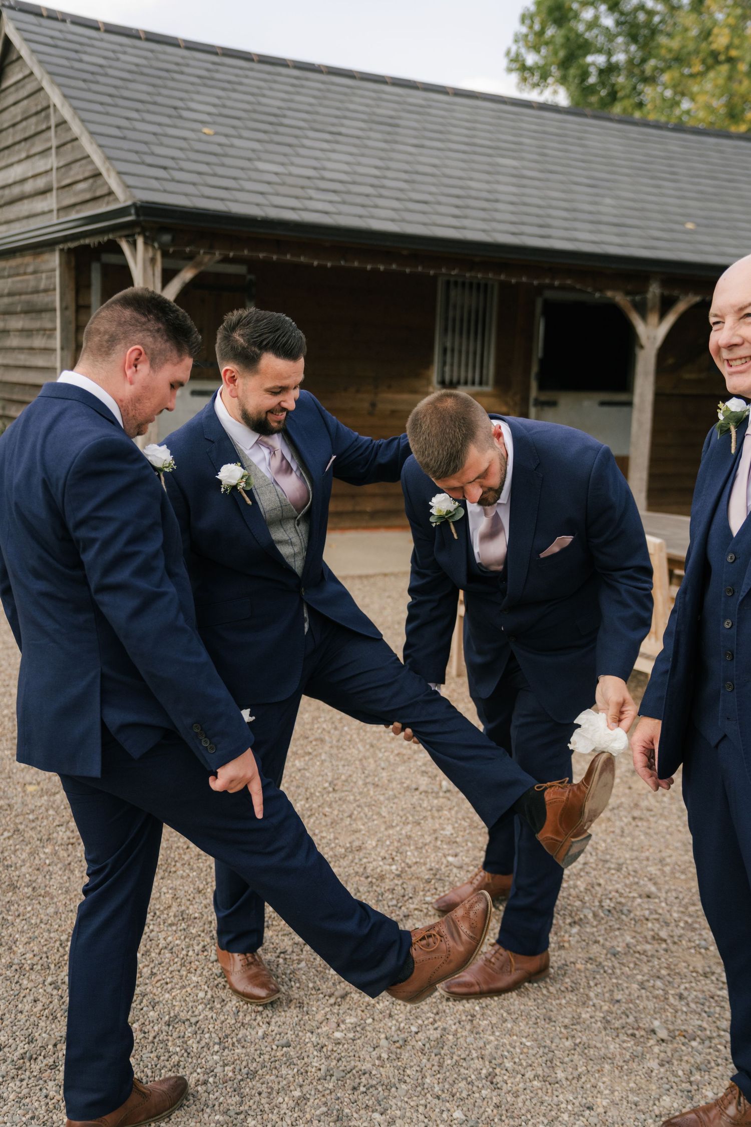 Groomsmen in navy suits inspect their brown leather dress shoes while standing in a line on gravel ground.