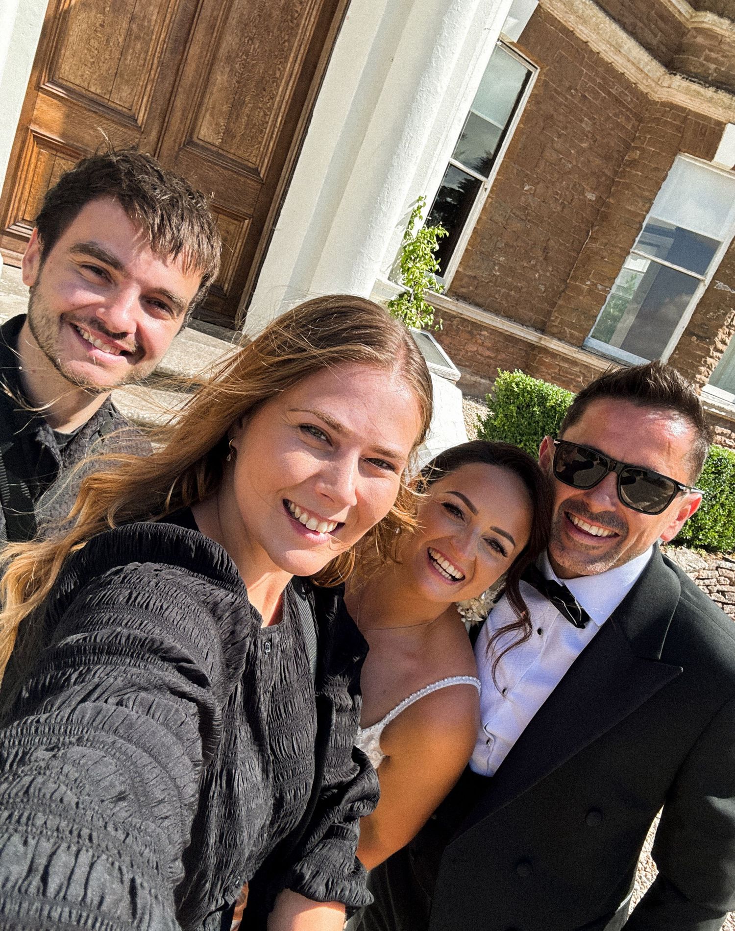 Four friends smiling together in a sunny selfie outside a brick building, dressed for a formal occasion.