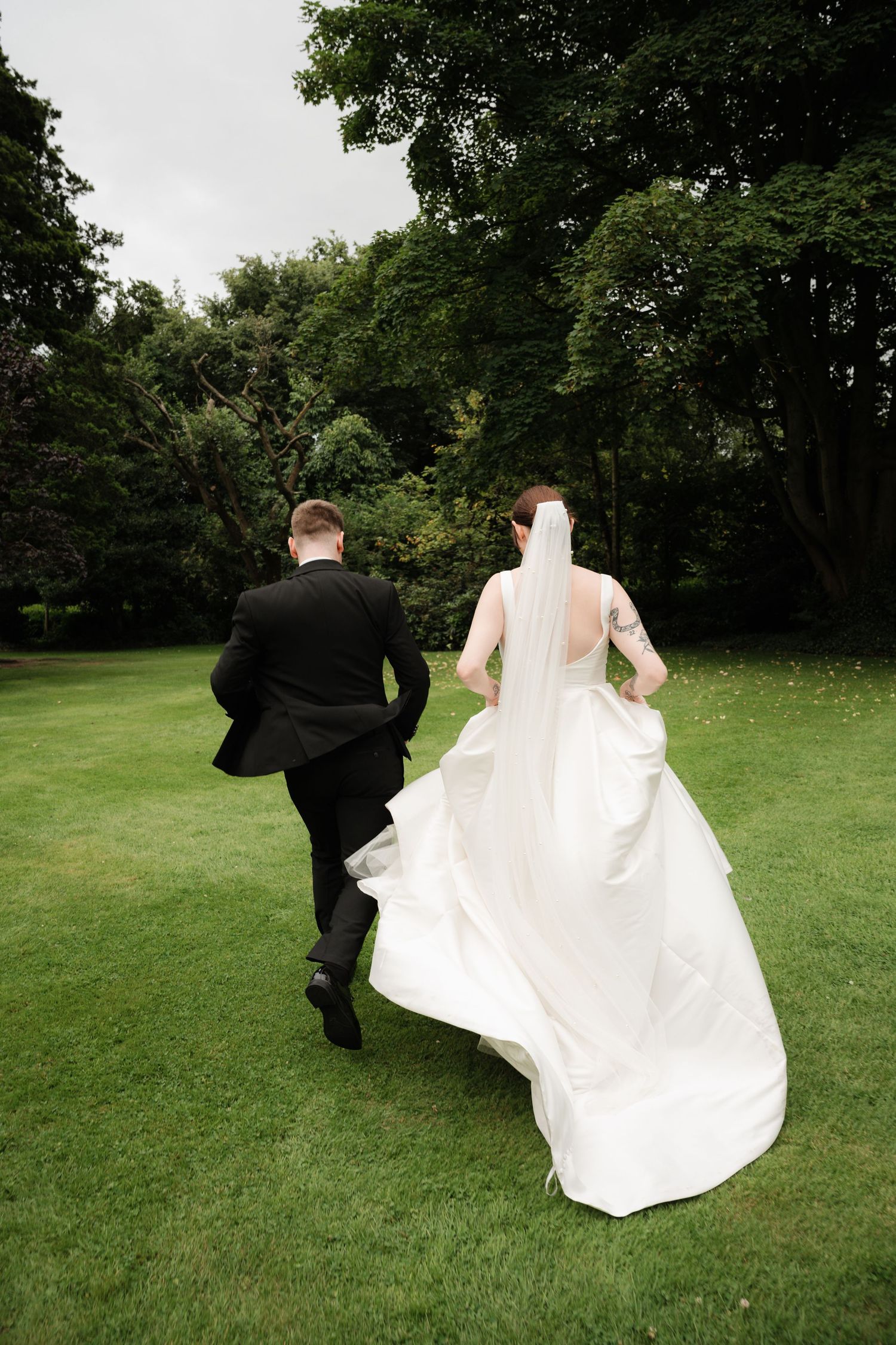 A couple in wedding attire runs playfully across a green lawn surrounded by trees in a sequence of action shots.