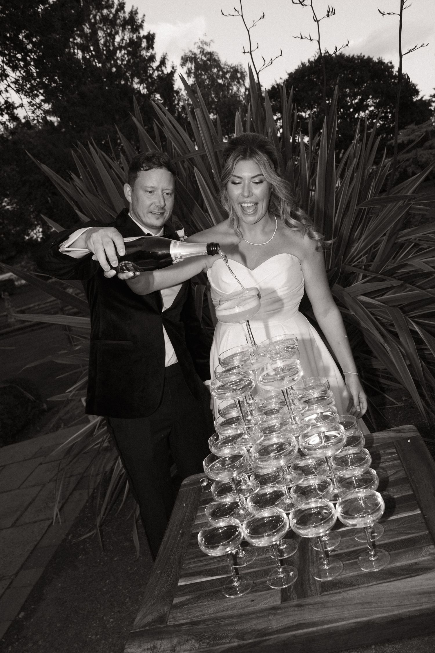 A couple carefully pours champagne into a tower of delicate coupe glasses at an elegant outdoor celebration.