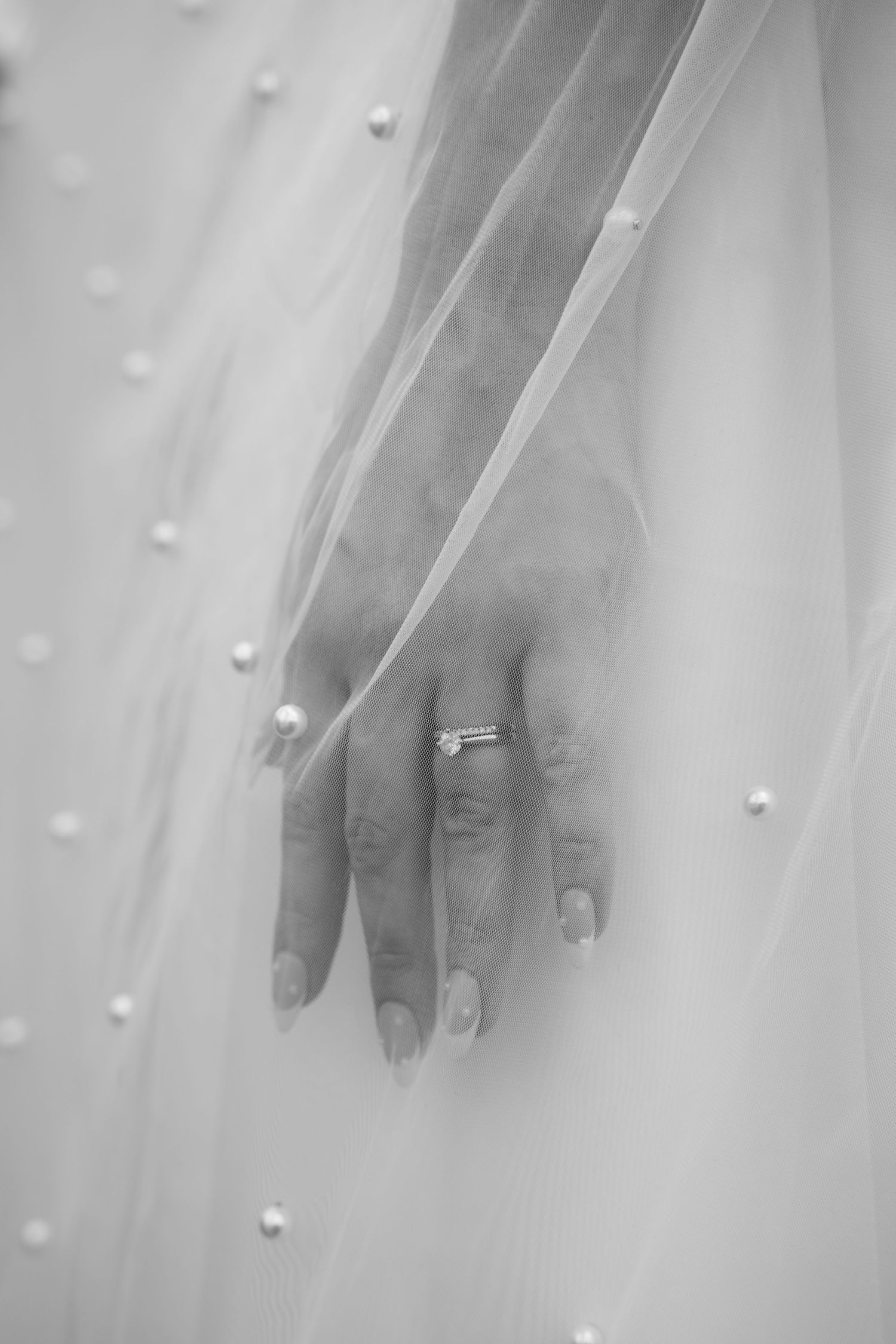 A hand wearing an engagement ring shows through delicate white tulle fabric with pearl details.