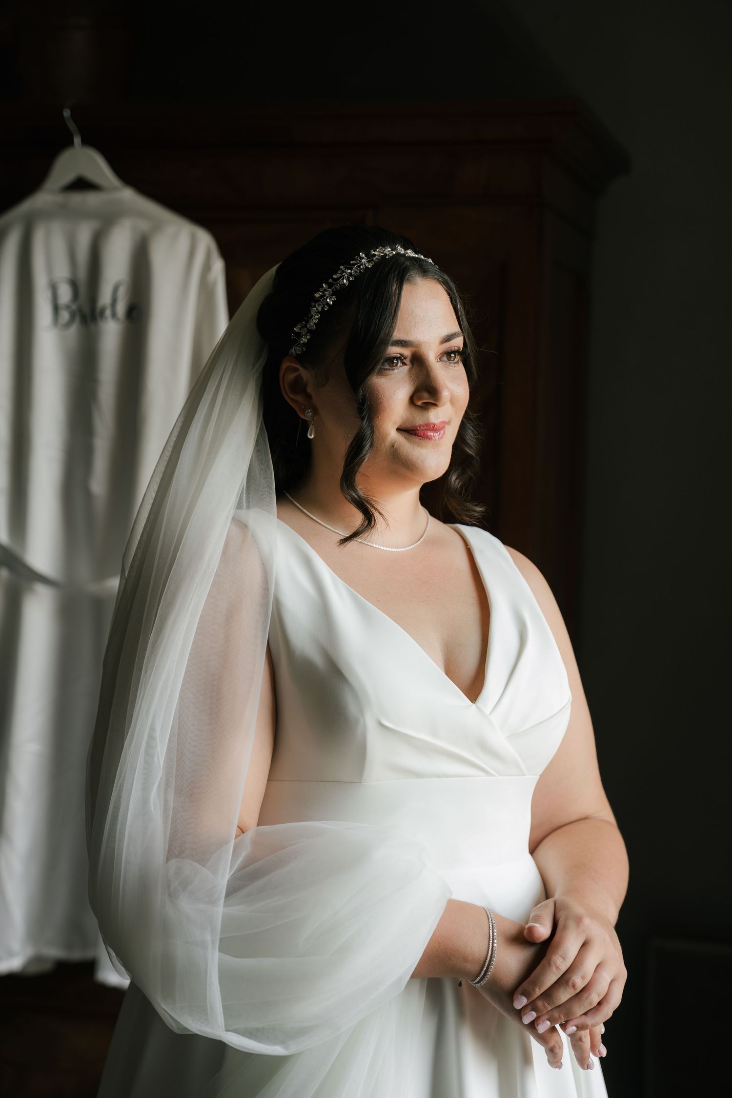 A bride in a white v-neck wedding dress poses near window light in a series of elegant bridal portraits.