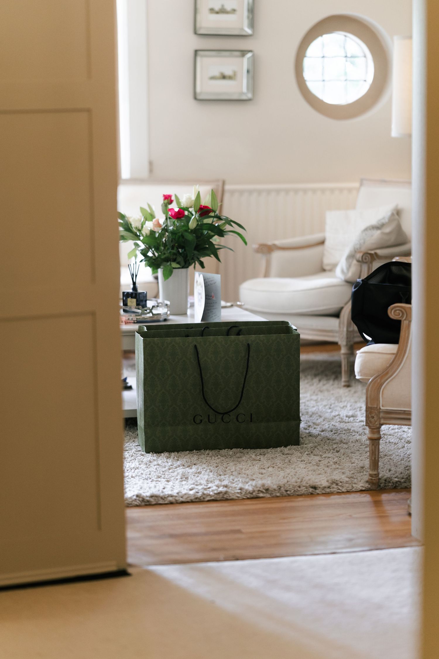 A cozy living room interior with a green Gucci shopping bag, white armchair, and pink flowers in a vase.