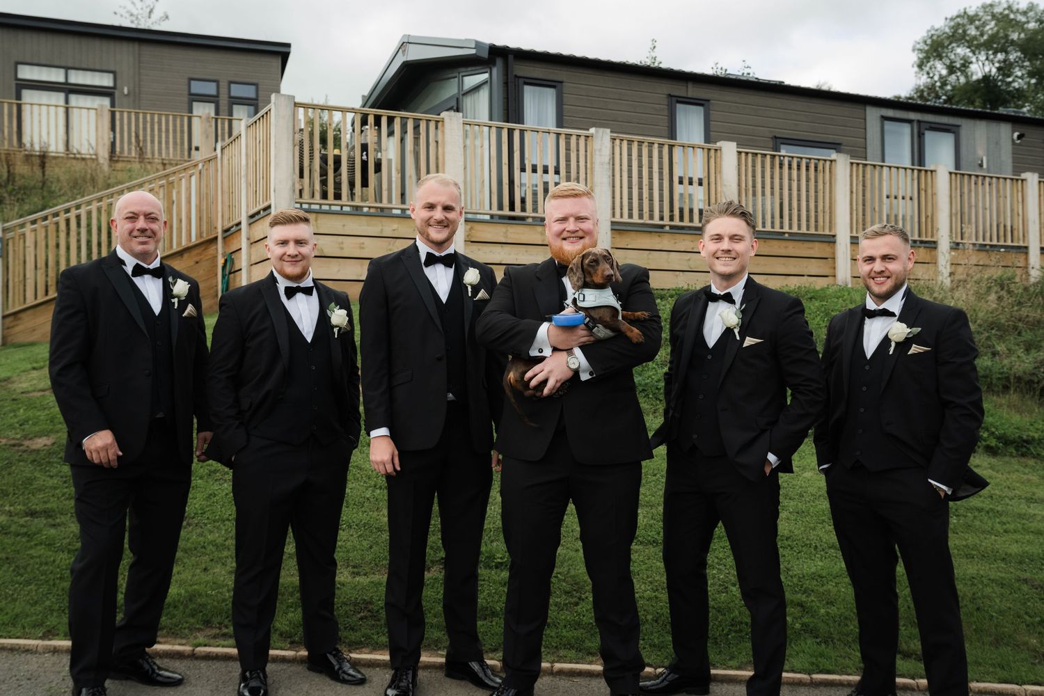 A wedding party in black tuxedos poses outdoors on a grassy hill with a wooden deck visible in the background.