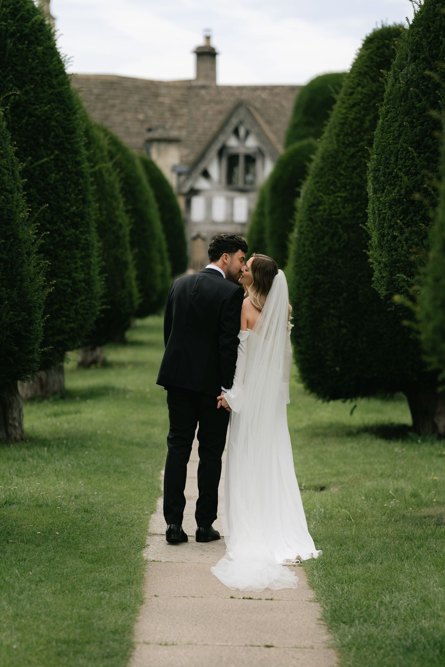 A couple in wedding attire walks along a path between perfectly manicured tall hedges in a formal garden setting.
