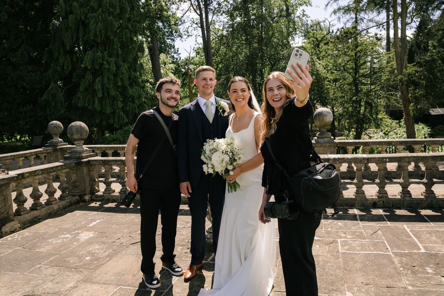 Wedding couple poses for selfie with guests on stone terrace surrounded by lush green trees.