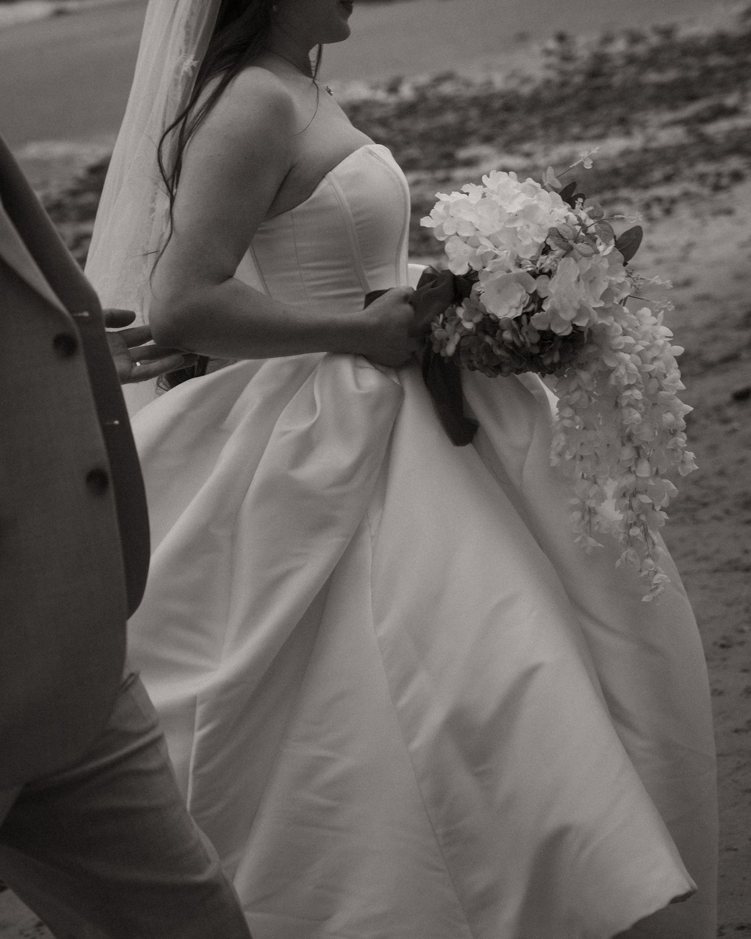 Black and white photo of a bride in a strapless gown holding a cascading floral bouquet while walking outdoors.