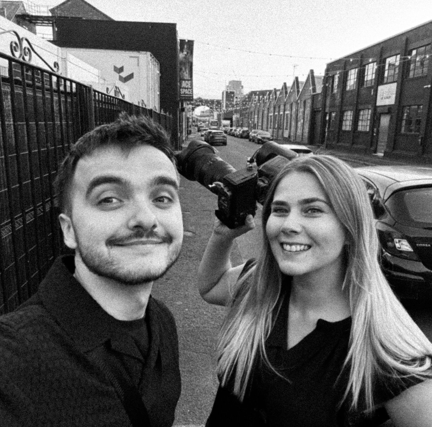 Black and white selfie of two people smiling together on a city street with buildings and parked cars in background.