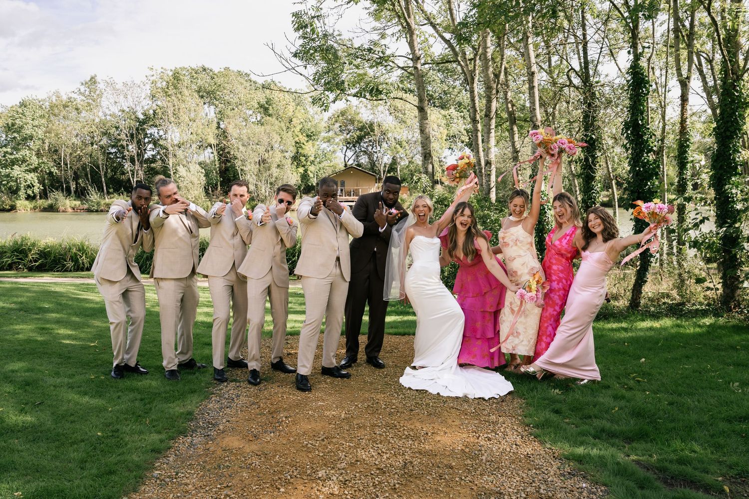 Wedding party celebrating outdoors, bride in white gown with bridesmaids in pink and groomsmen in tan suits, trees in background.