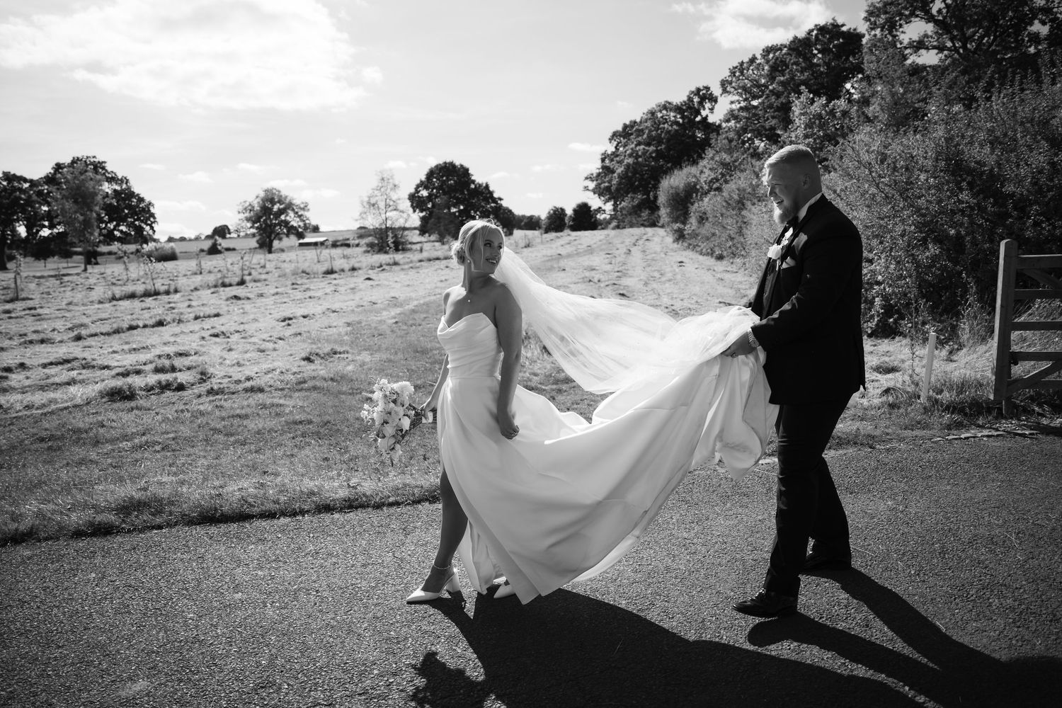 A couple in wedding attire walks through a scenic outdoor setting as a flowing veil catches the wind.