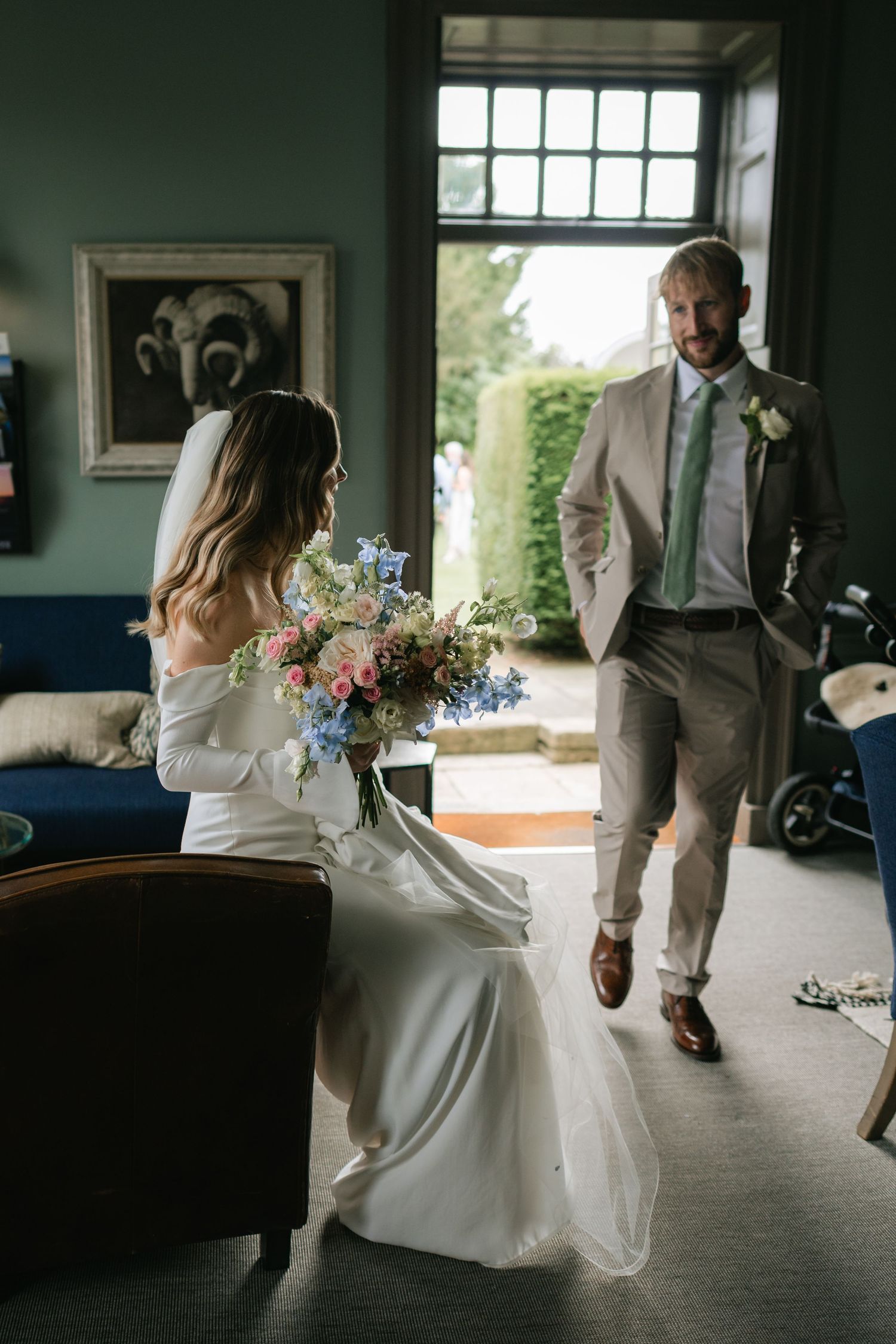 A bride in a white off-shoulder gown holds a colorful bouquet while seated as a groom in a light gray suit stands nearby.