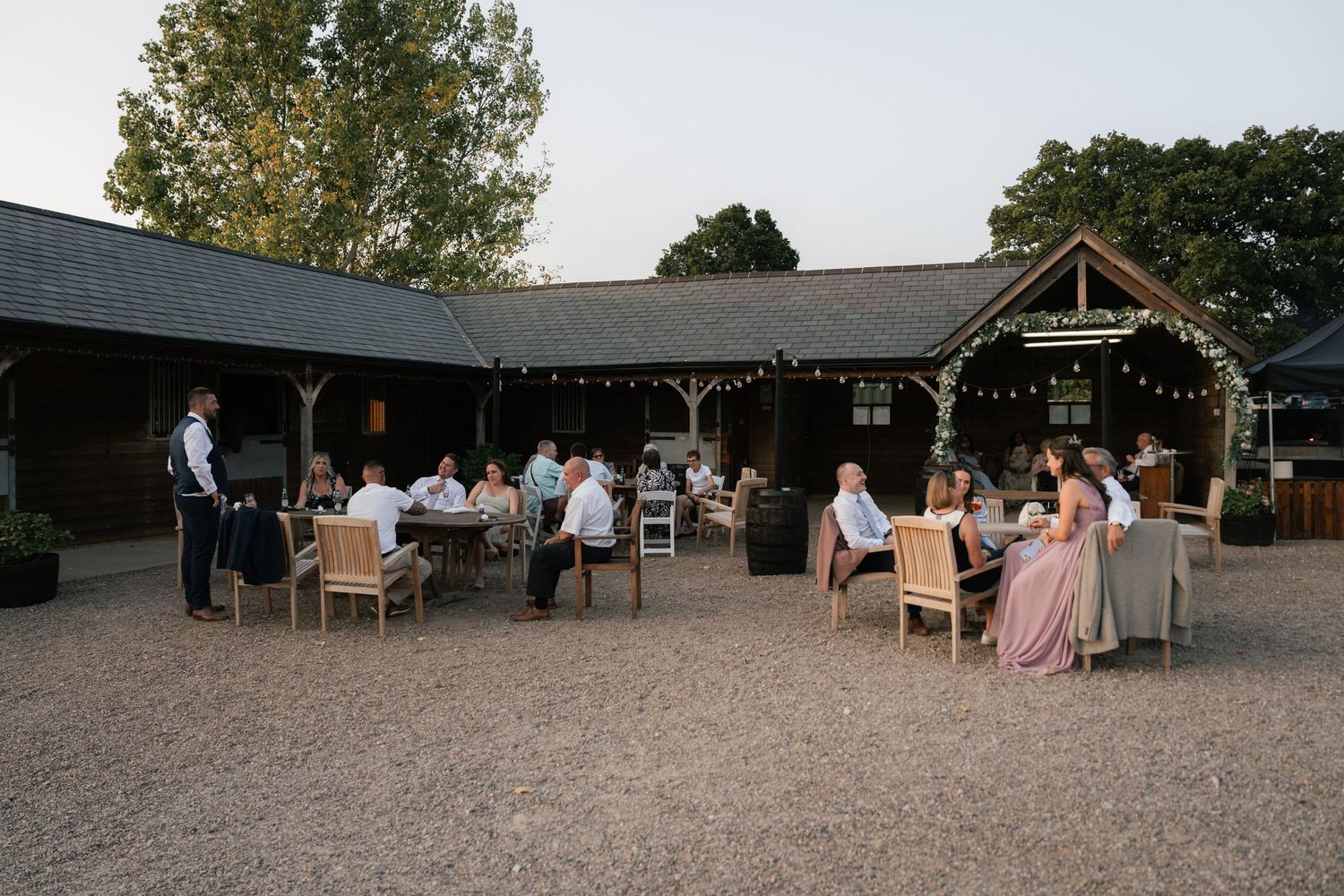 Panoramic view of a rustic outdoor venue with guests gathered for a celebration under trees and wooden structures.