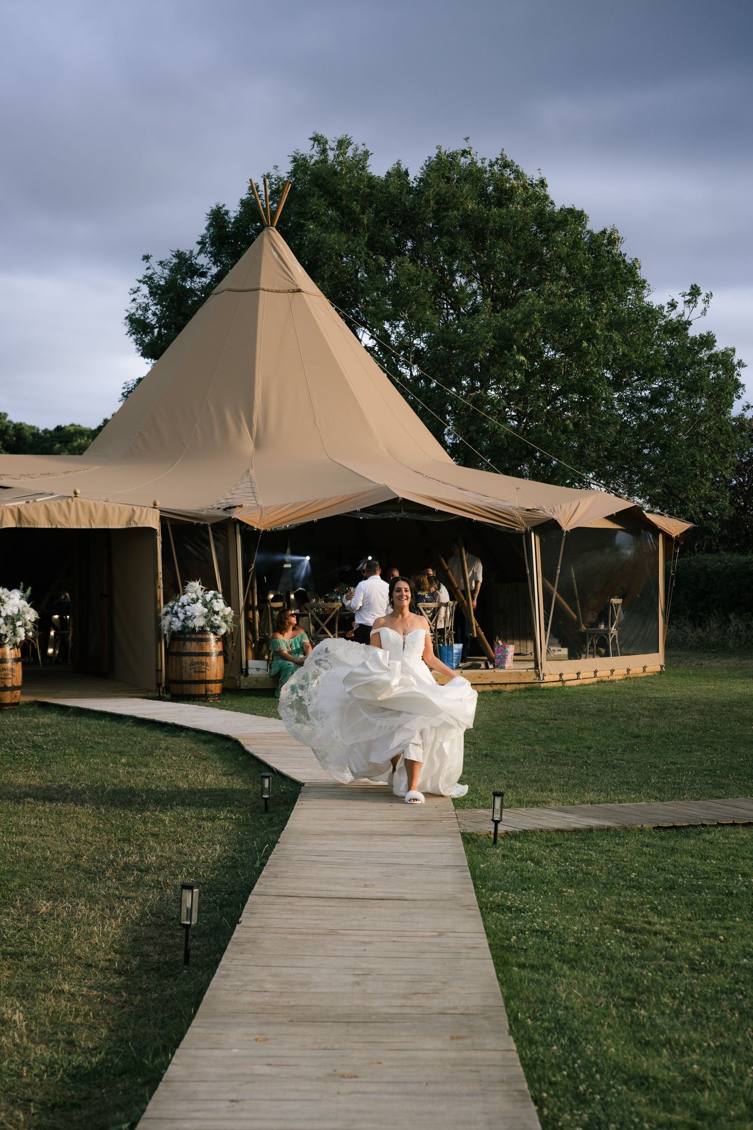 A series of tan outdoor event tents connected by a wooden walkway on a grassy field during a wedding celebration.
