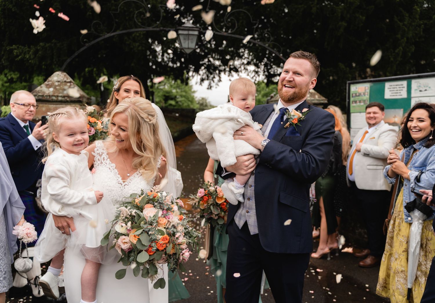 A joyful wedding celebration outdoors with confetti flying as people gather for photos with floral bouquets.