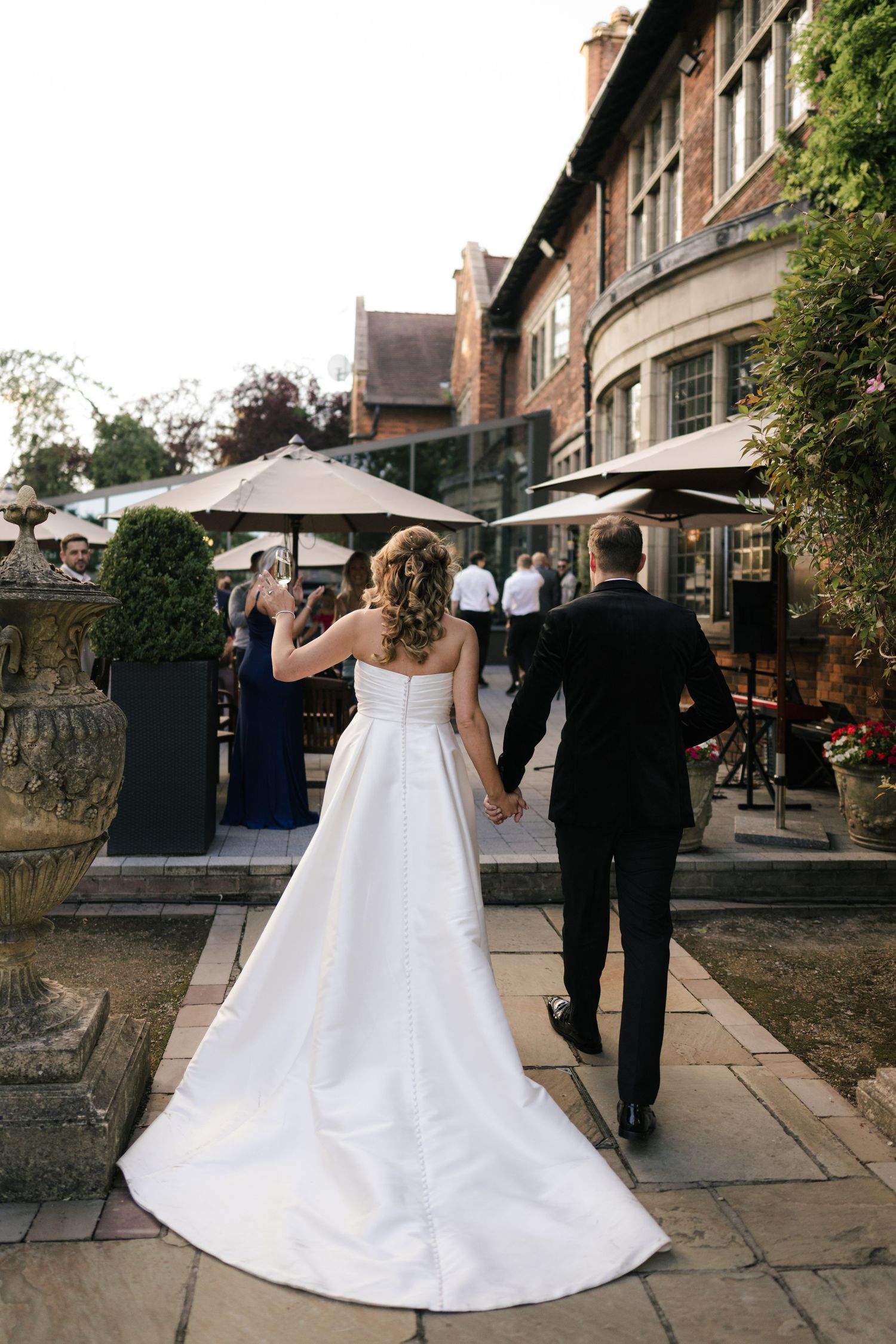 A couple walks along a stone path outside a historic brick building, with the bride wearing a flowing white gown.