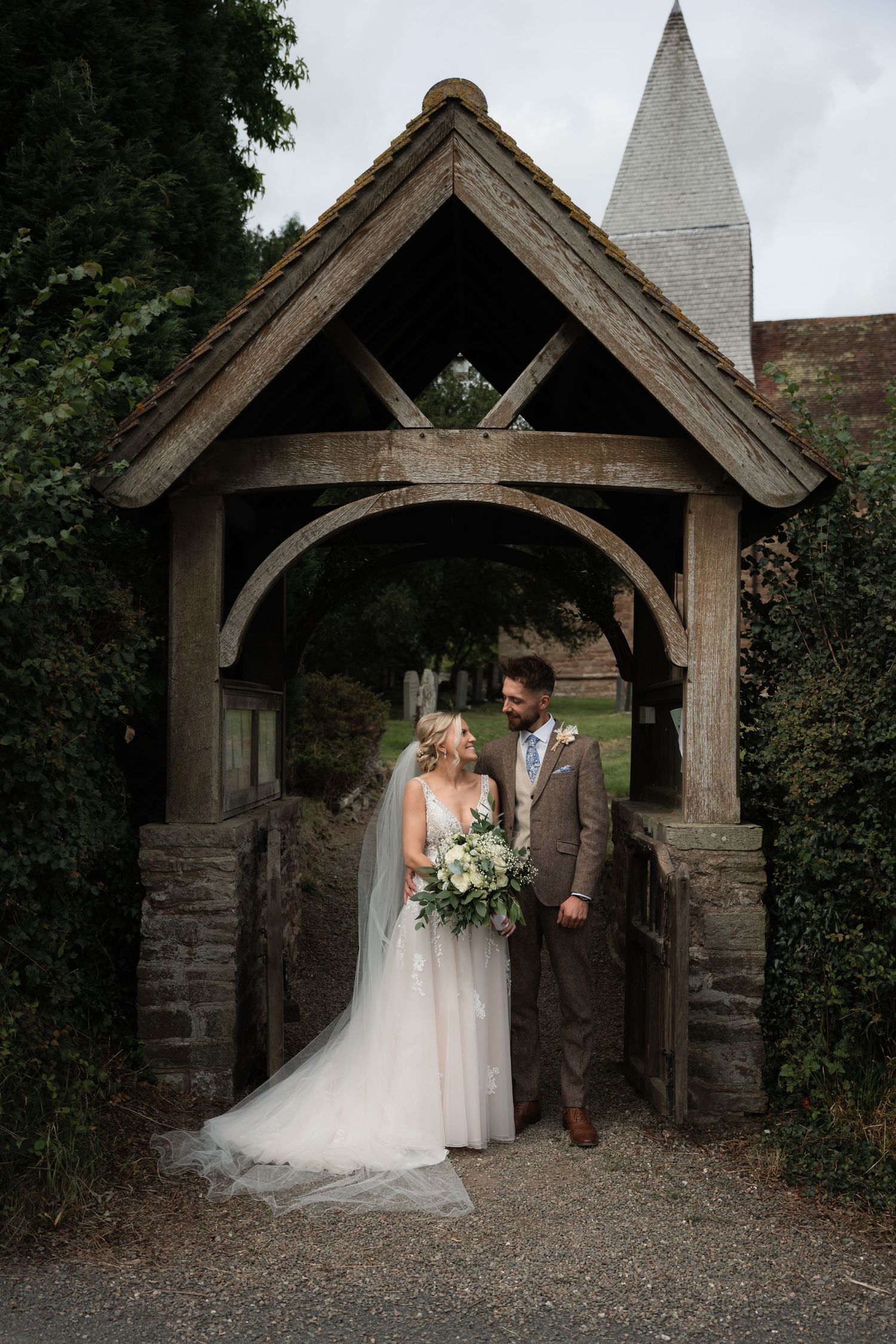 Couple shares intimate moments under rustic wooden church lychgate during their countryside wedding celebration.