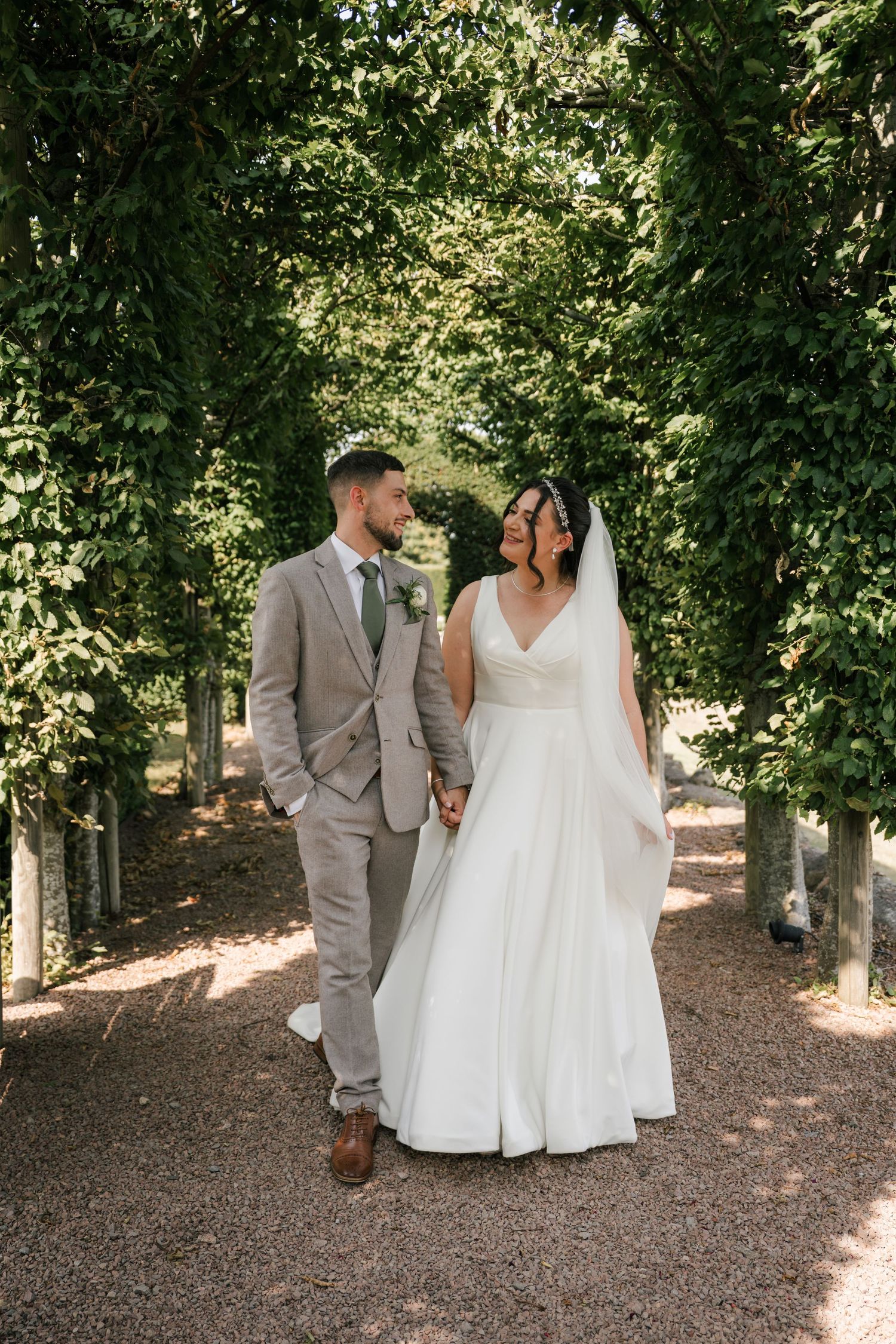 A couple in wedding attire walks hand in hand down a tree-lined garden path on their special day.