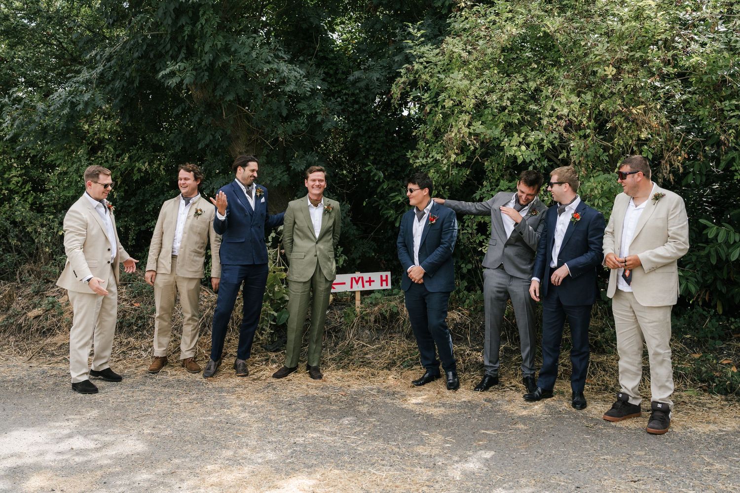 A group of groomsmen in various colored suits stand together on a path outdoors with greenery in the background.