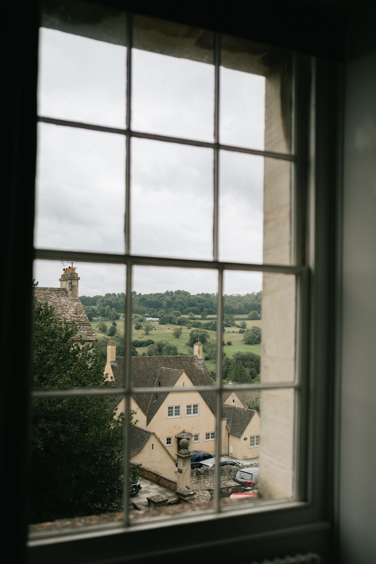 View through a window overlooking a quaint village with stone houses and rolling hills in the countryside.