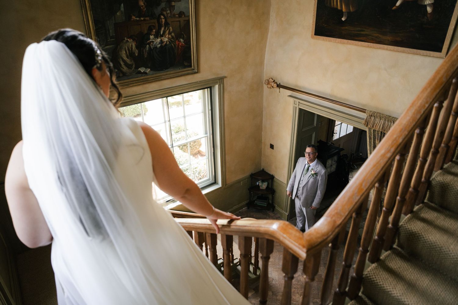 A bride in a white wedding dress and veil walks up a wooden staircase in a sunlit room.