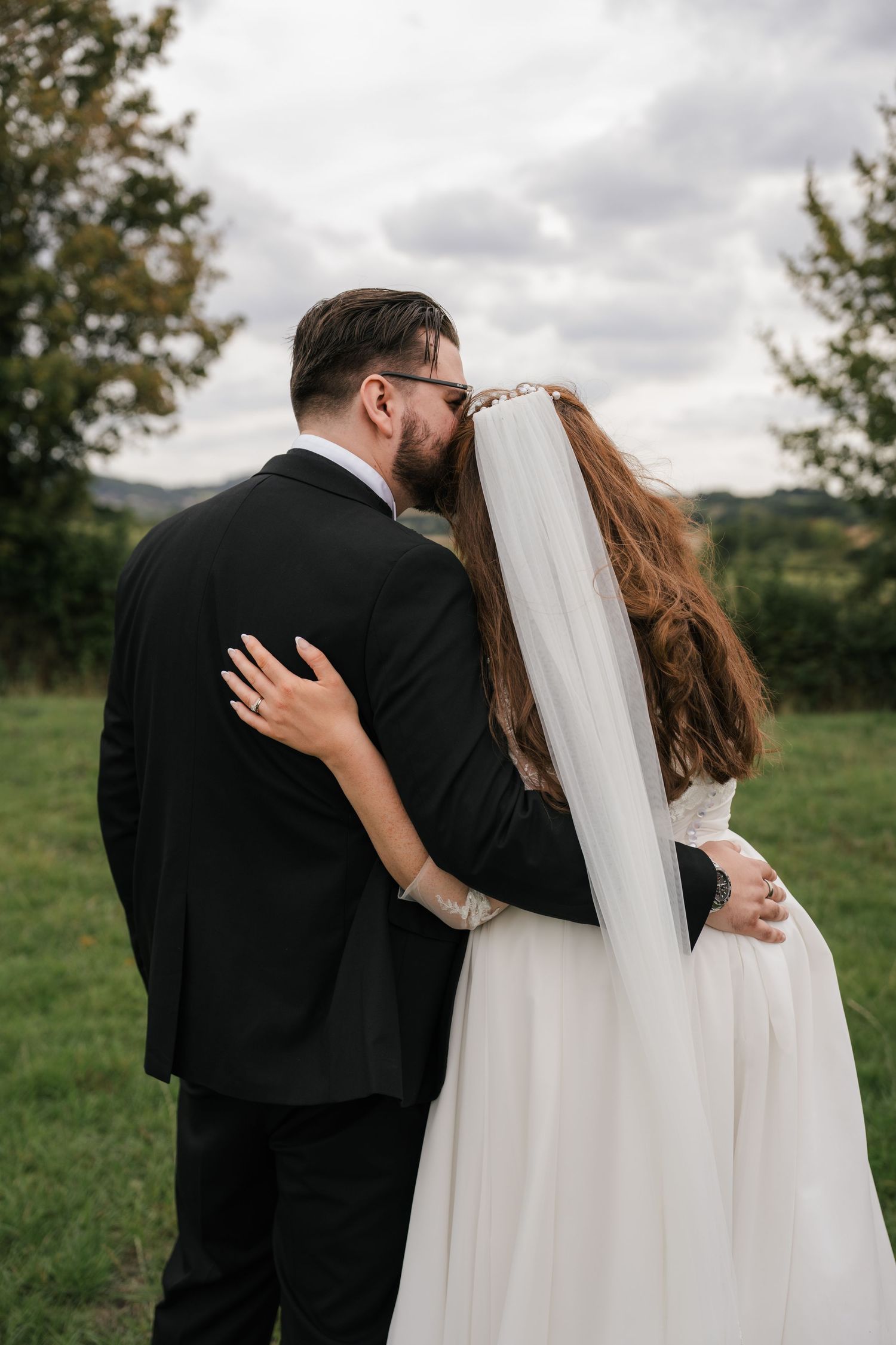 A romantic wedding couple embraces in a grassy field under cloudy skies.