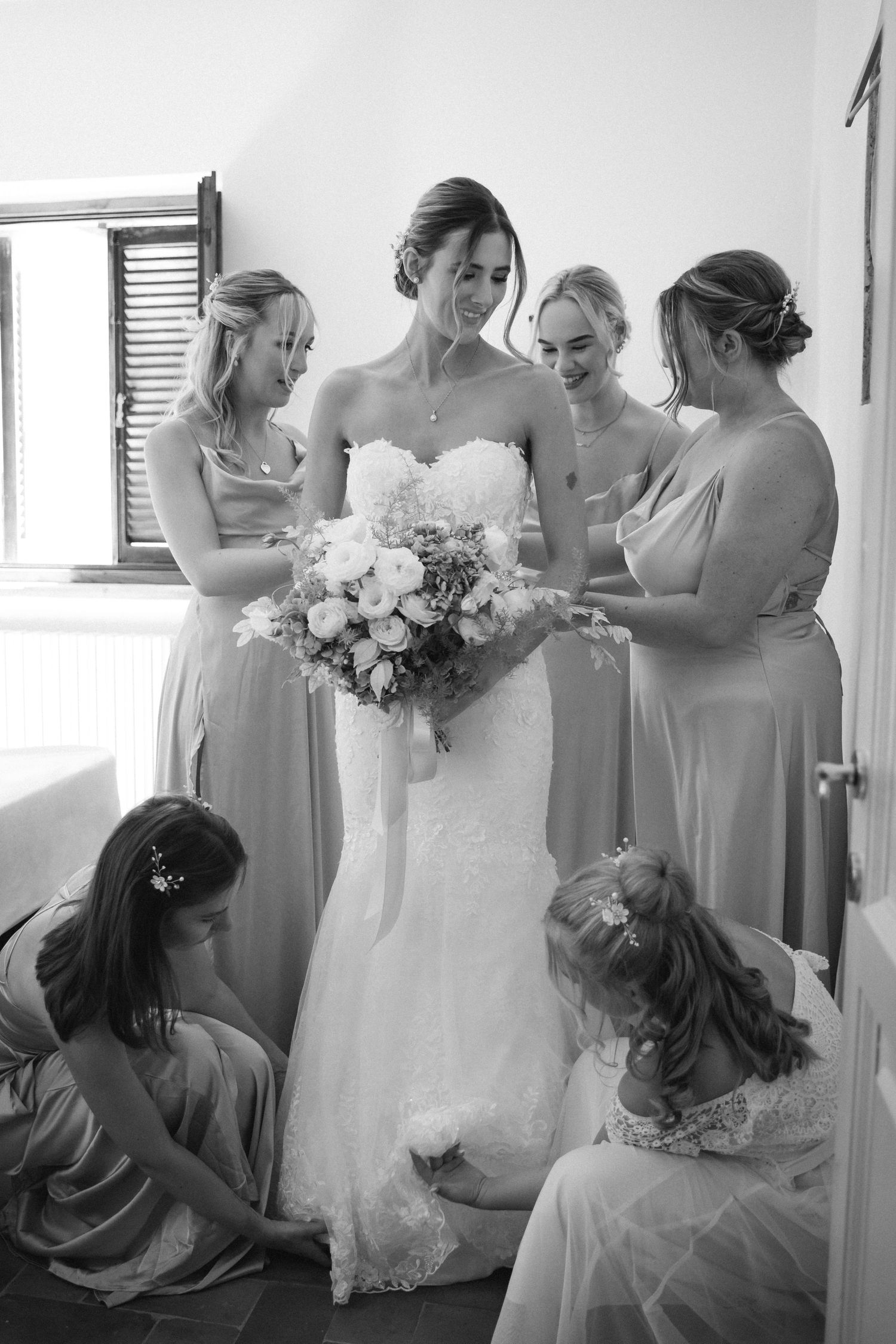 Black and white image showing bridesmaids helping with wedding preparations in a sunlit room.