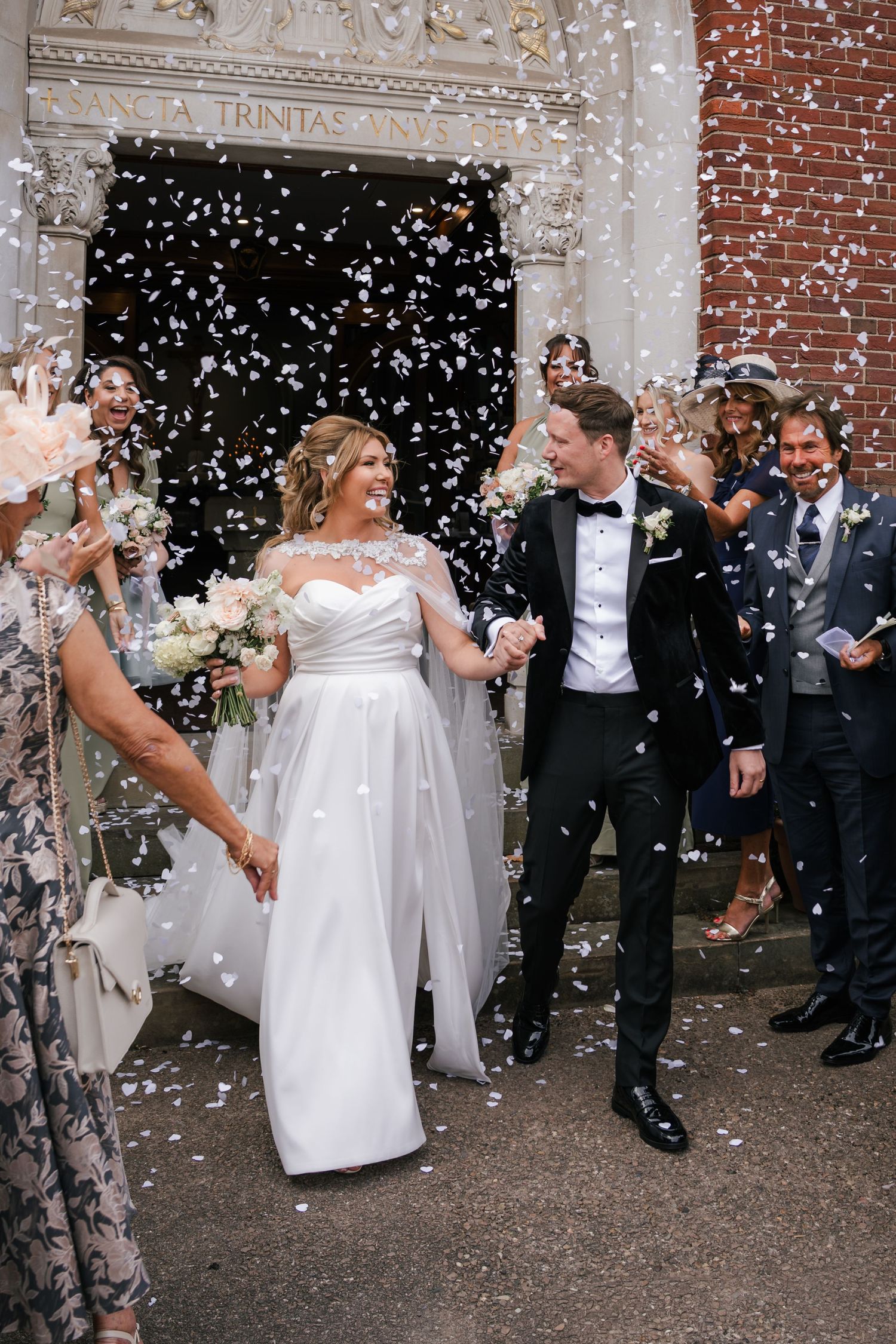 A wedding celebration outside a brick church building with guests throwing confetti as newlyweds celebrate their special moment.