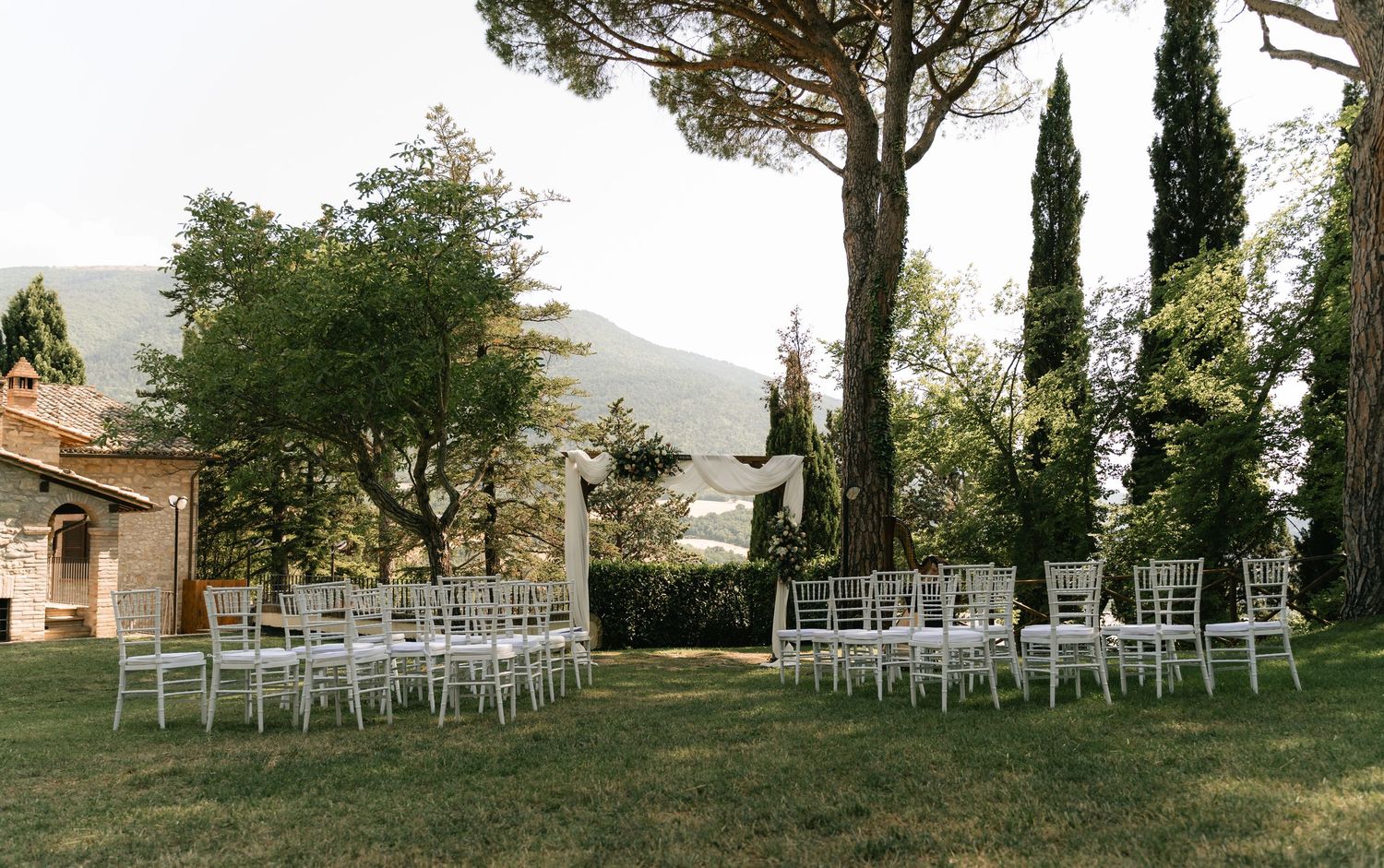 Row of white ceremony chairs arranged outdoors among cypress trees against mountain backdrop.
