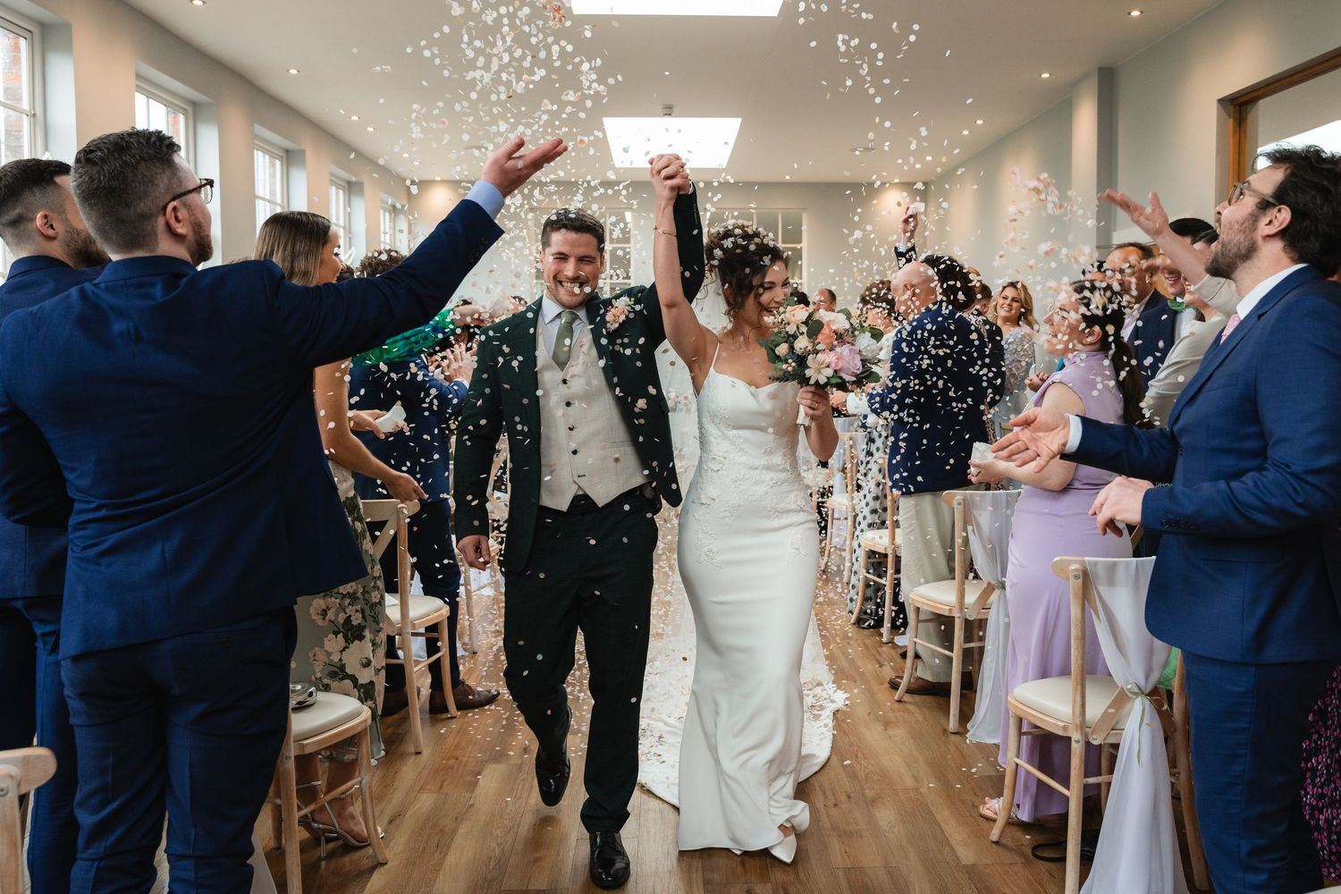 Wedding guests shower a couple with confetti as they walk down a bright hallway during a joyous celebration.