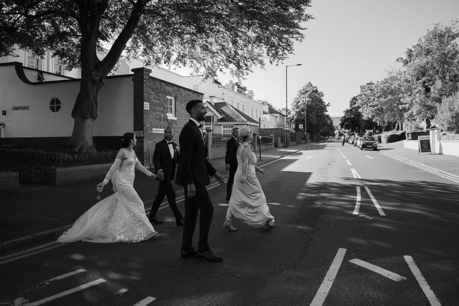 A wedding party walks along a tree-lined street while a photographer captures candid moments in black and white.