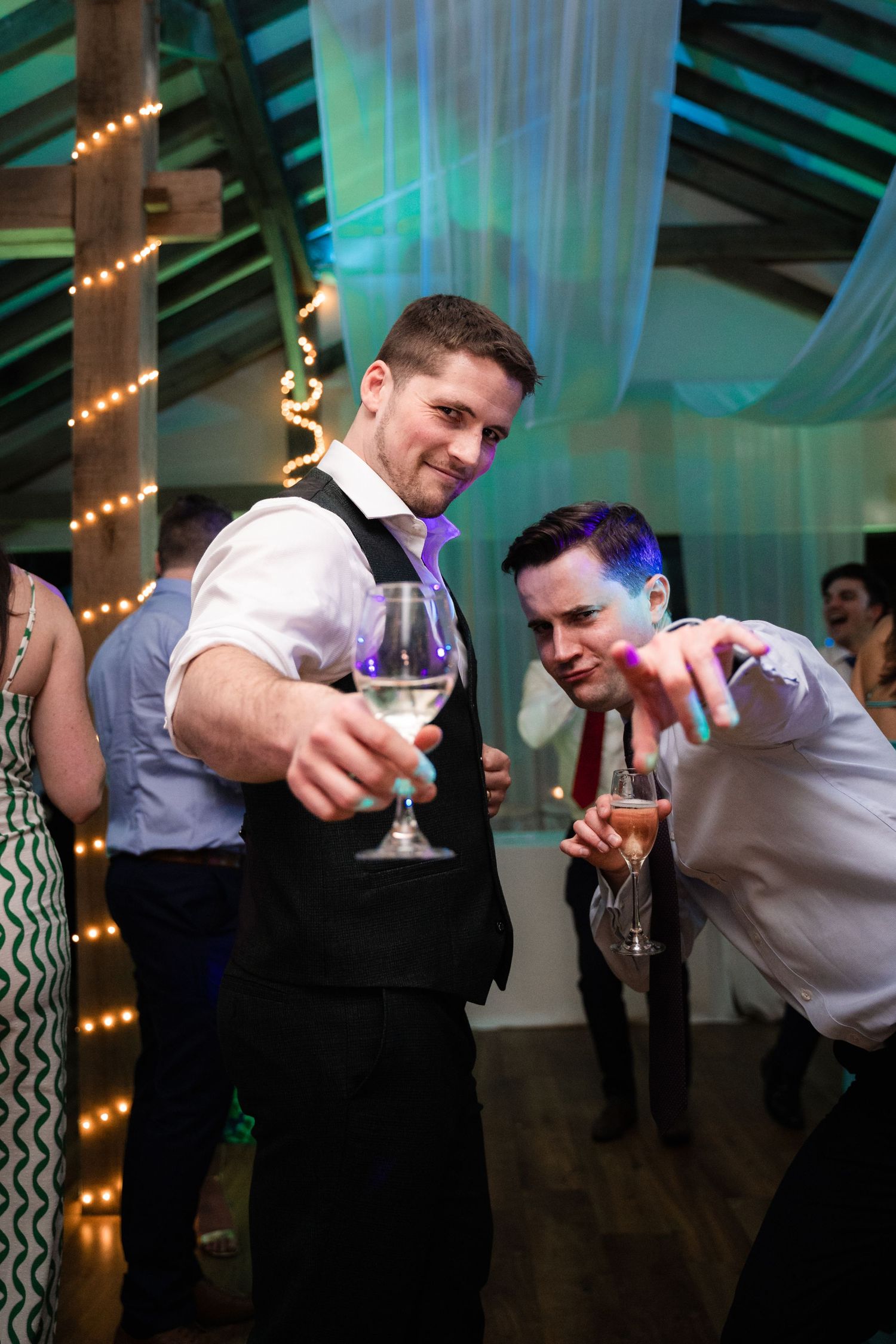 Two people in formal attire holding cocktail glasses at an event with teal lighting and string lights in the background.