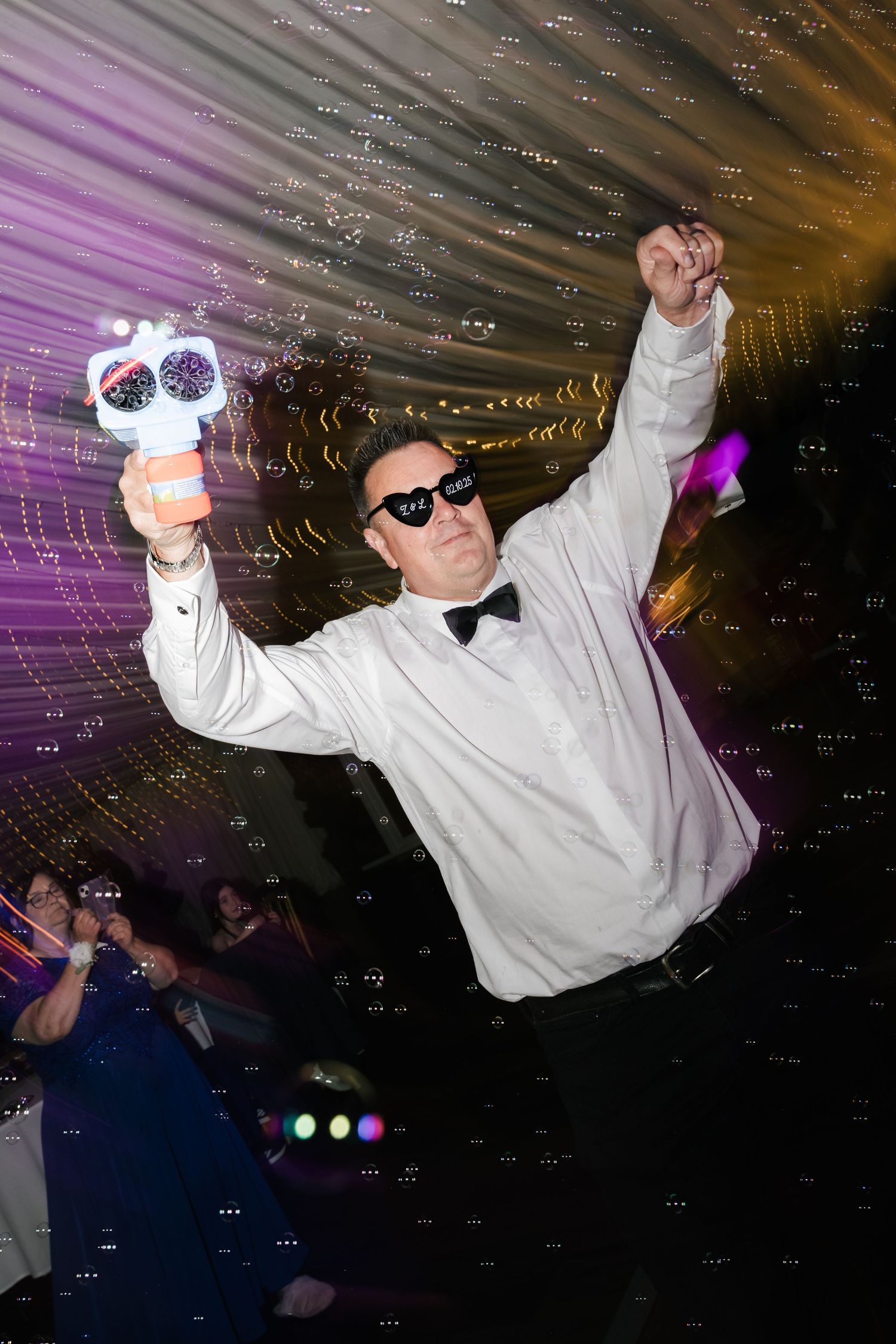 Person in white shirt and bowtie dancing energetically at a party with colorful lights in the background.