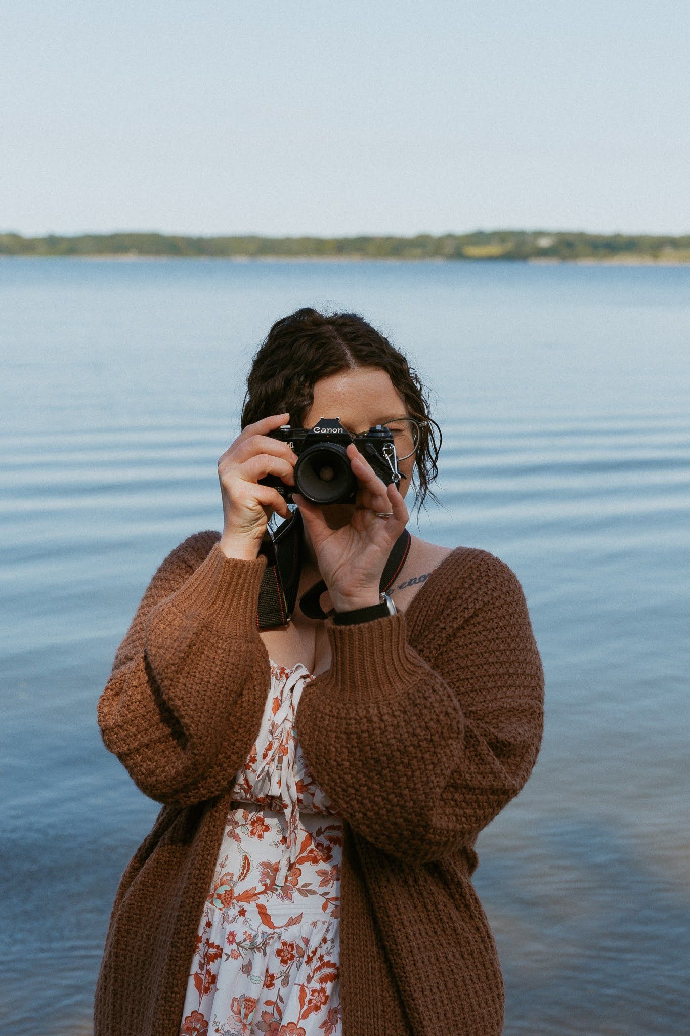 A photographer wearing a brown knit cardigan and floral dress takes a picture by a calm lake on an overcast day.