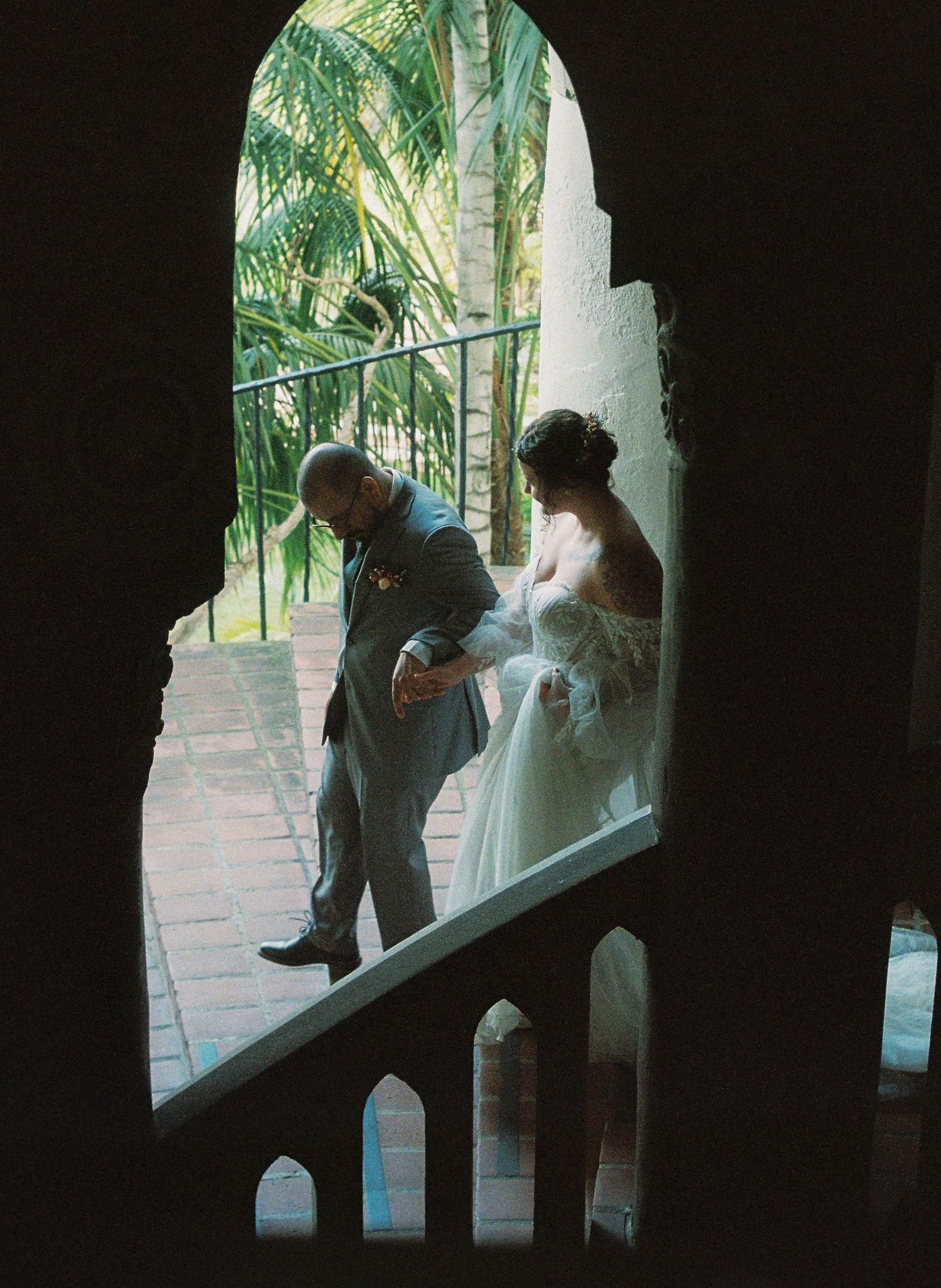 Two silhouettes stand in a window frame surrounded by palm trees in a romantic scene at dusk.