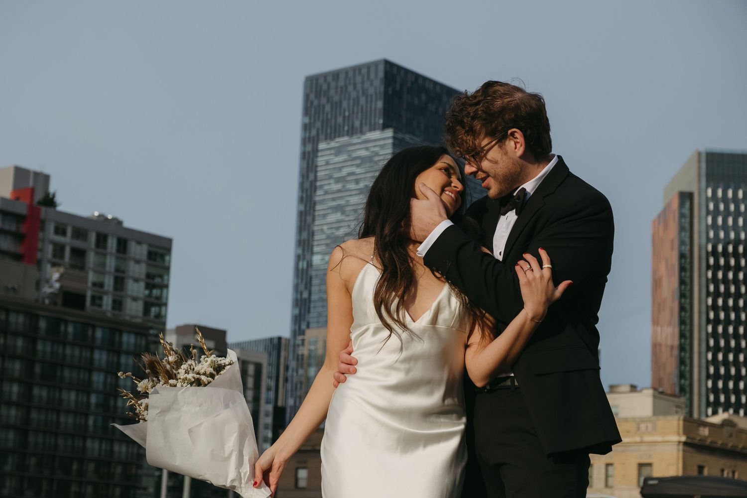 A bride and groom share a romantic moment on a rooftop with city skyscrapers in the background on a sunny day.
