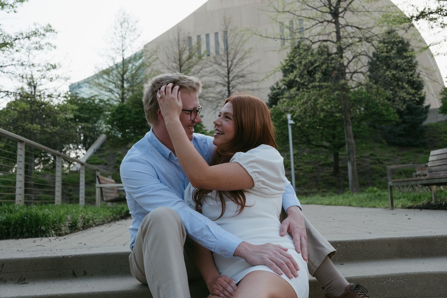 Couple sitting on outdoor steps sharing a romantic moment, smiling and embracing near a modern building.