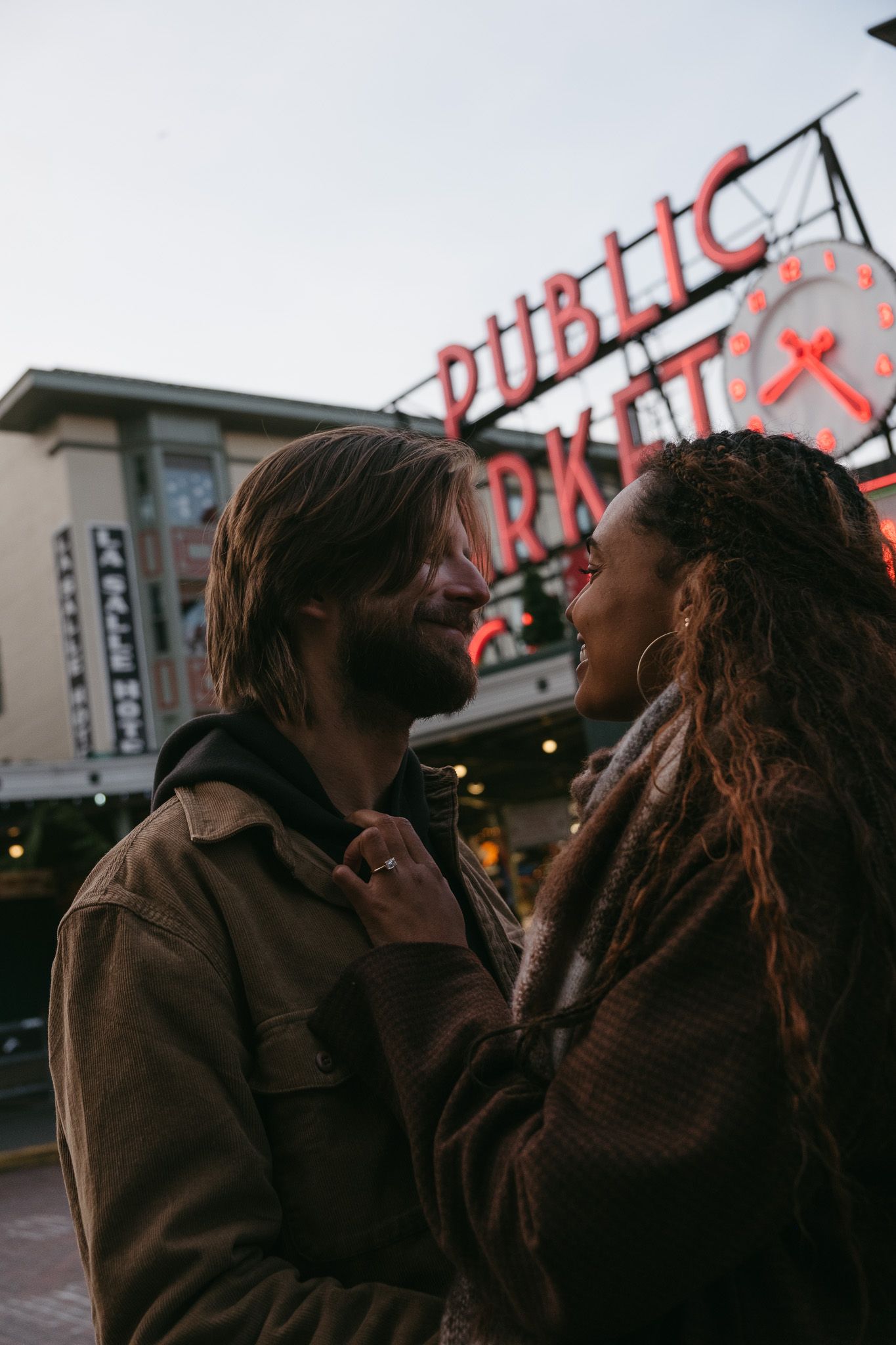 A couple embraces in a romantic moment outside a building with red signage at dusk.
