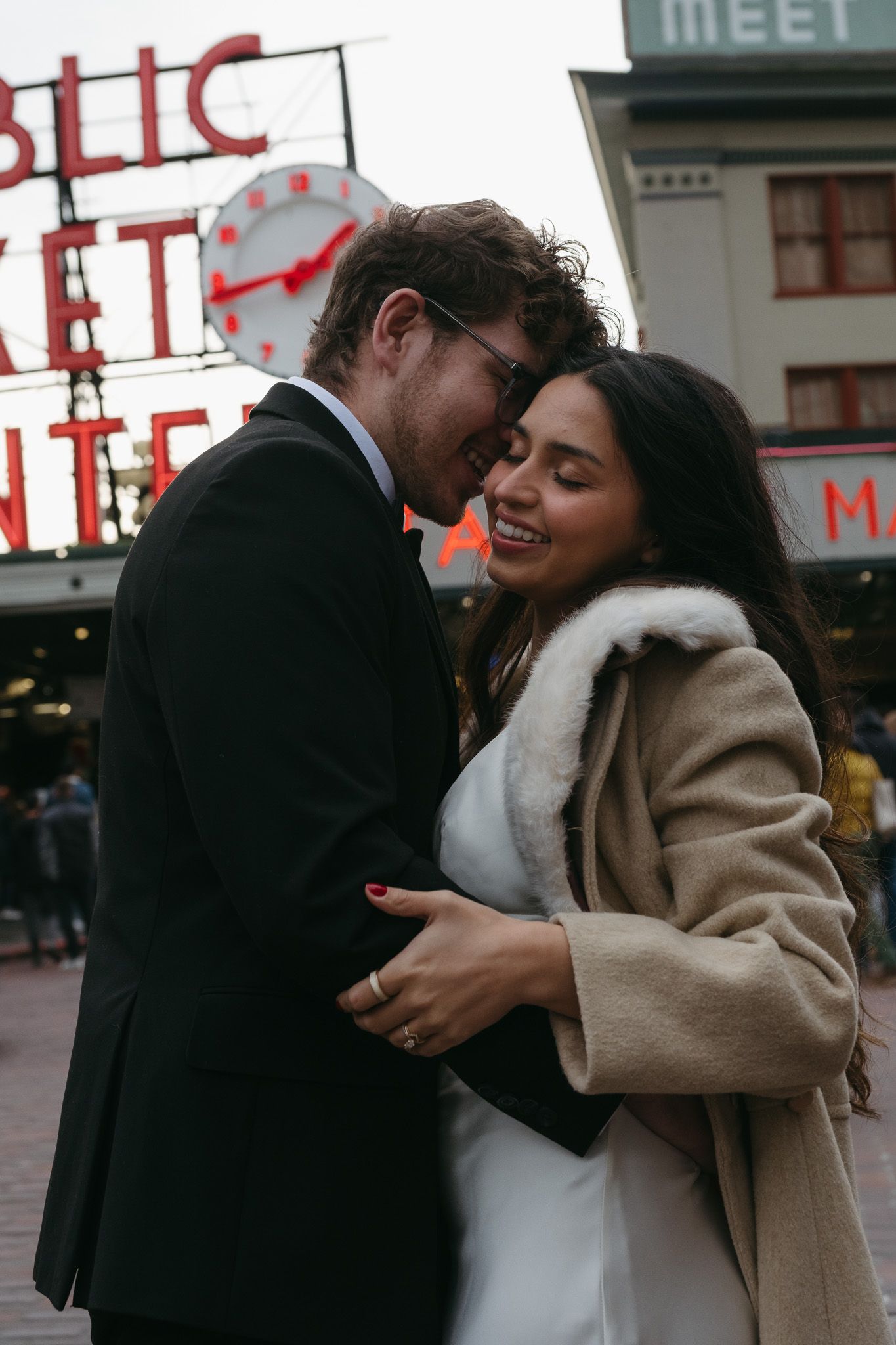 Couple embracing and smiling in front of Pike Place Market sign in Seattle, bride in white dress and fur coat.