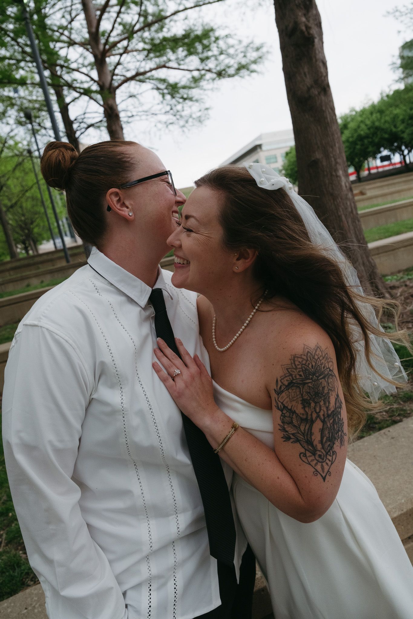 Two brides share a joyful moment, one in a white dress with veil and tattoo, the other in a white shirt and tie.