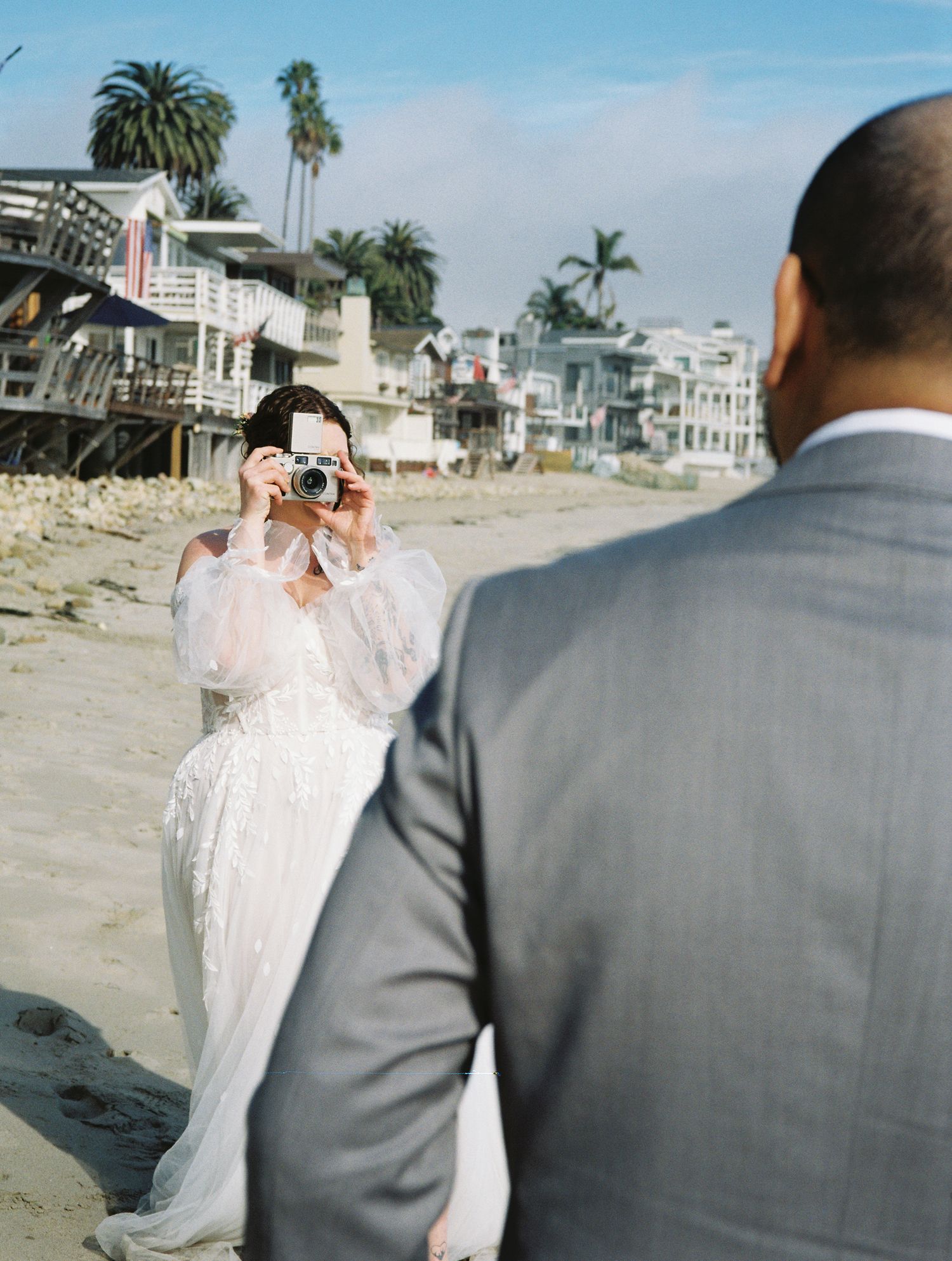 A couple shares a romantic moment on a sunny beach with palm trees and coastal homes in the background.