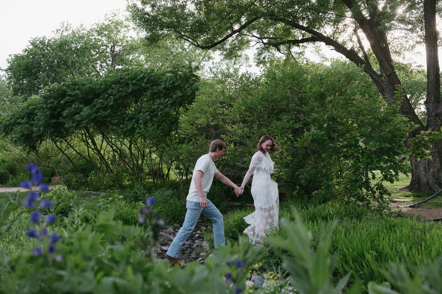 Couple holding hands walking through a lush green garden with blooming flowers and trees.