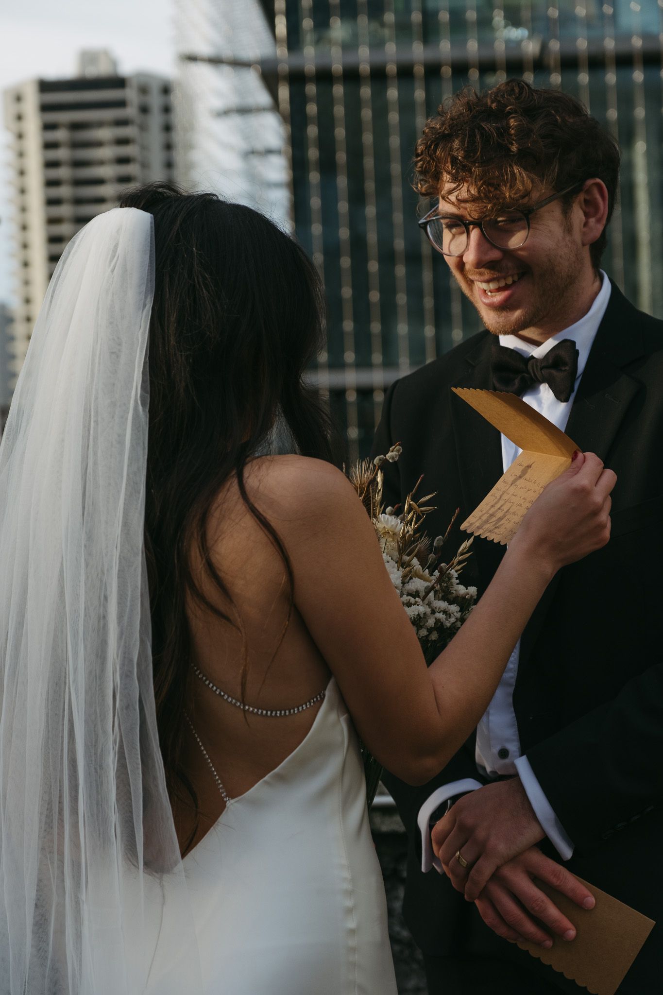 Bride in white gown and veil shares vows with smiling groom in tuxedo on rooftop with city skyline backdrop.