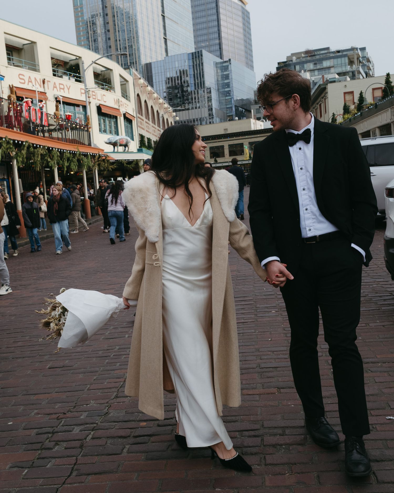 A couple in wedding attire walks along a brick paved area near city buildings.