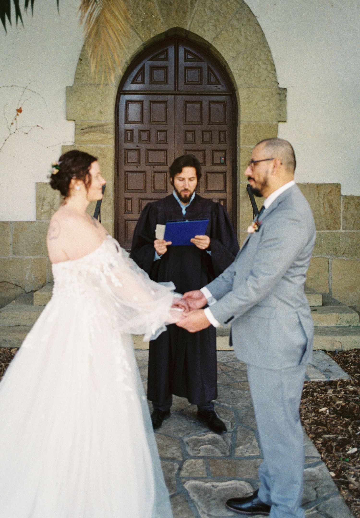 A wedding ceremony takes place in front of a grand arched wooden door at a stone church.